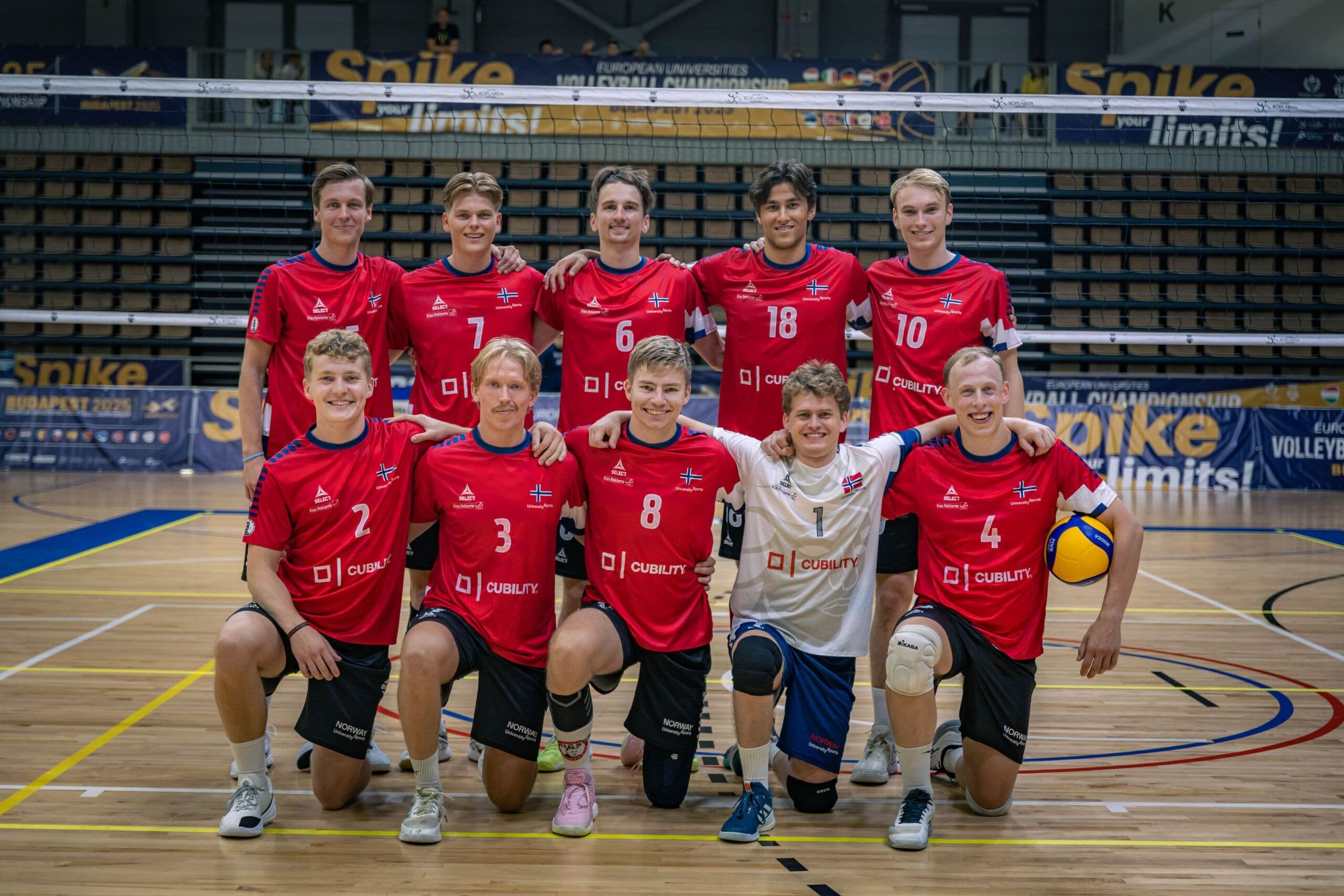Et mannlig volleyballag i røde drakter poserer samlet på banen i en idrettshall.