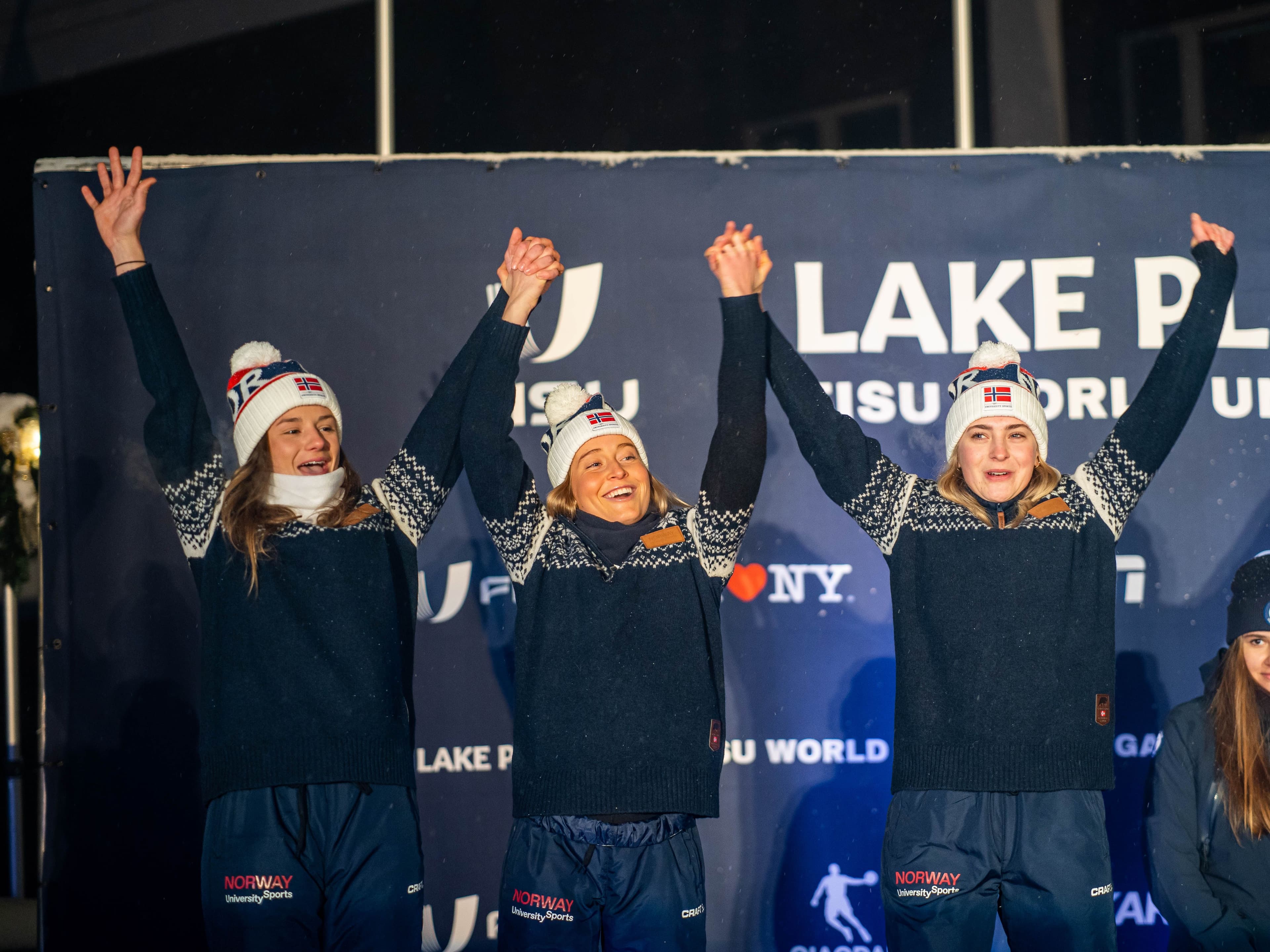 Tre kvinnelige langrennsutøvere i norske landslagsklær jubler med hevede armer på seierspallen under FISU World University Games i Lake Placid.
