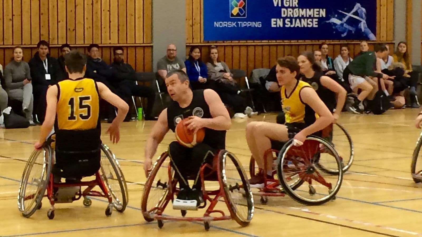 Rullestolbasketkamp i en idrettshall der en spiller holder ballen mellom to motspillere i gule drakter.