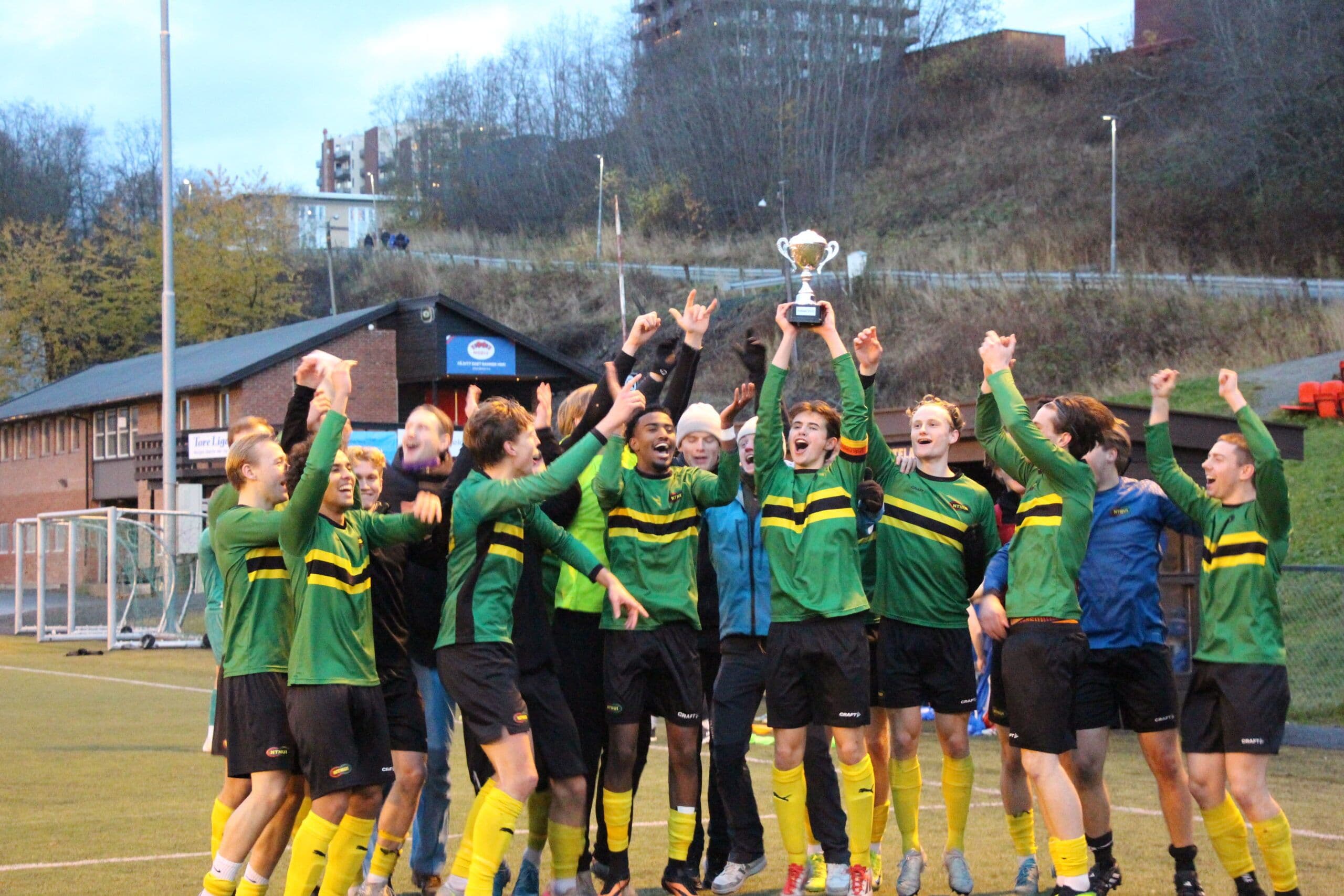 Et fotballag i grønt og gult jubler og løfter en pokal på en kunstgressbane.