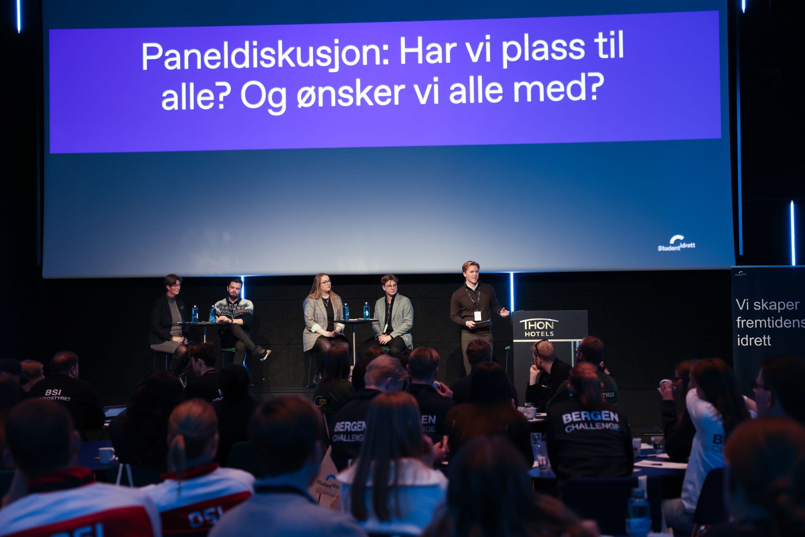 Fem personer i en paneldebatt på en scene foran publikum, under skjermteksten «Paneldiskusjon: Har vi plass til alle? Og ønsker vi alle med?».