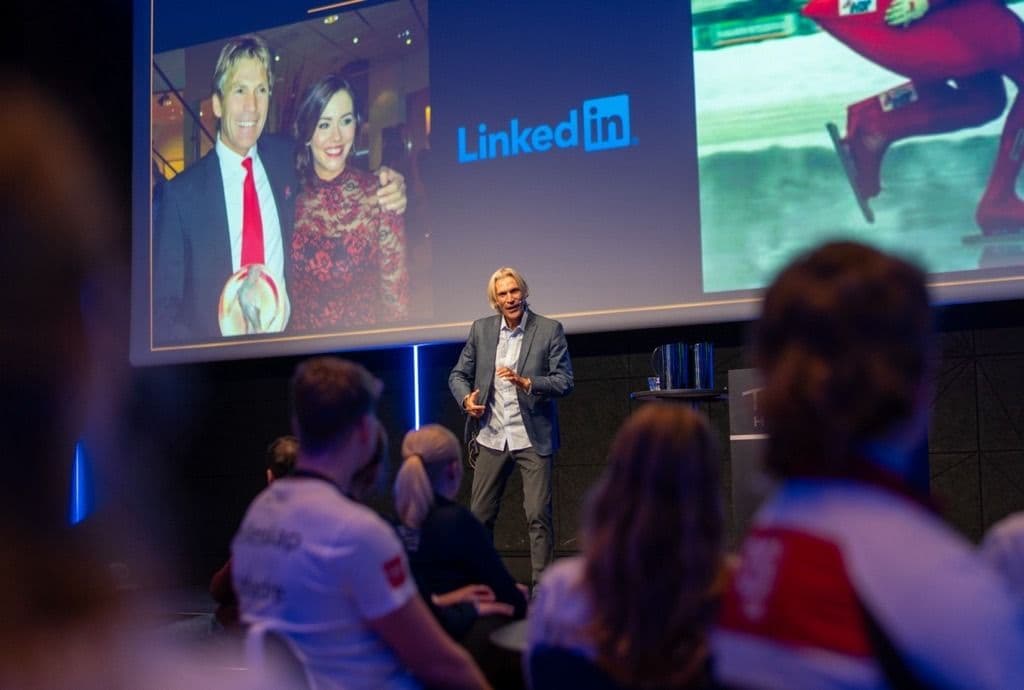 En foredragsholder står på scenen foran publikum med en LinkedIn-presentasjon på storskjermen bak seg.