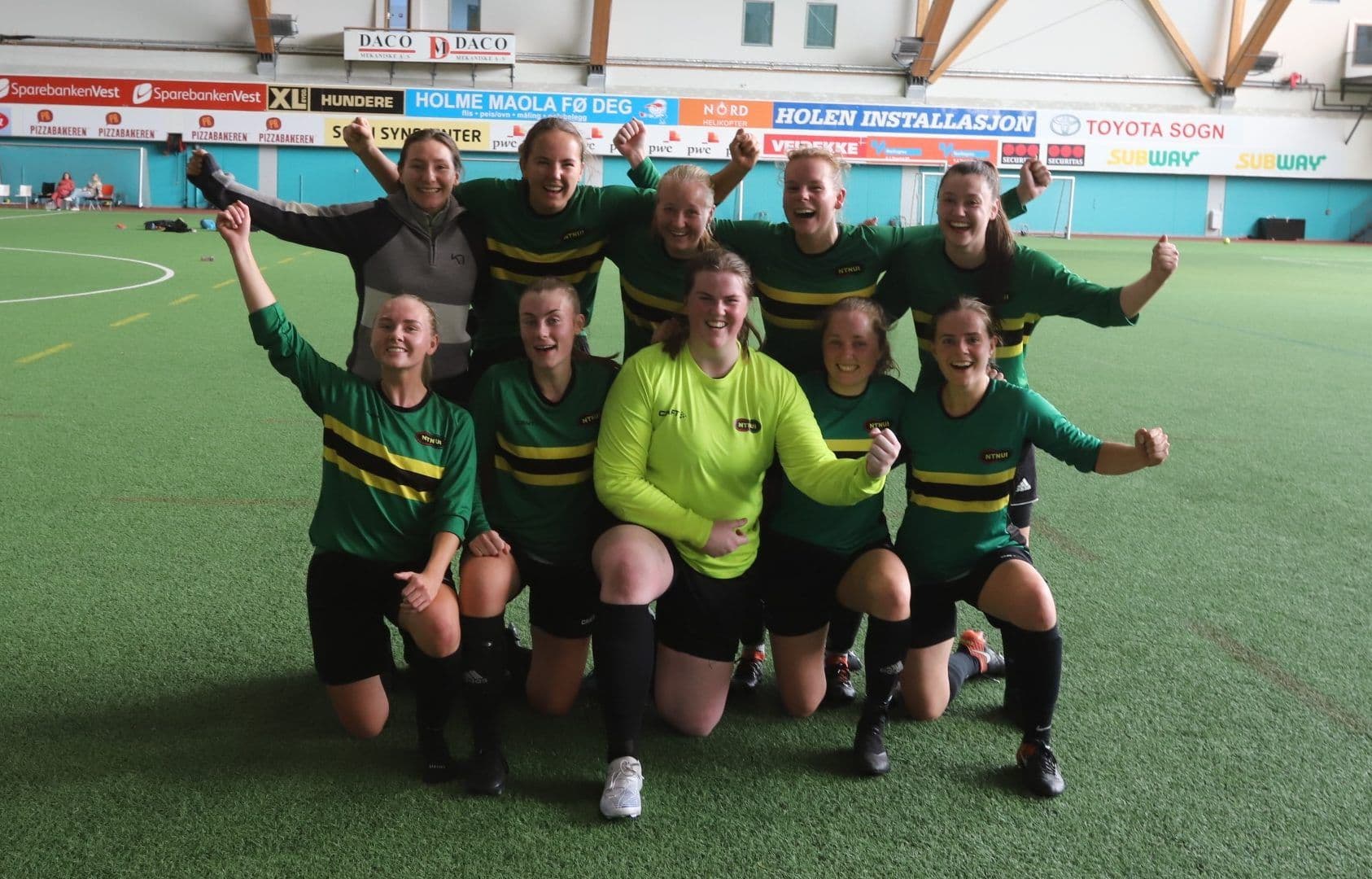 Et kvinnelig fotballag i grønne drakter og gul keeperdrakt jubler samlet i en innendørs fotballhall.