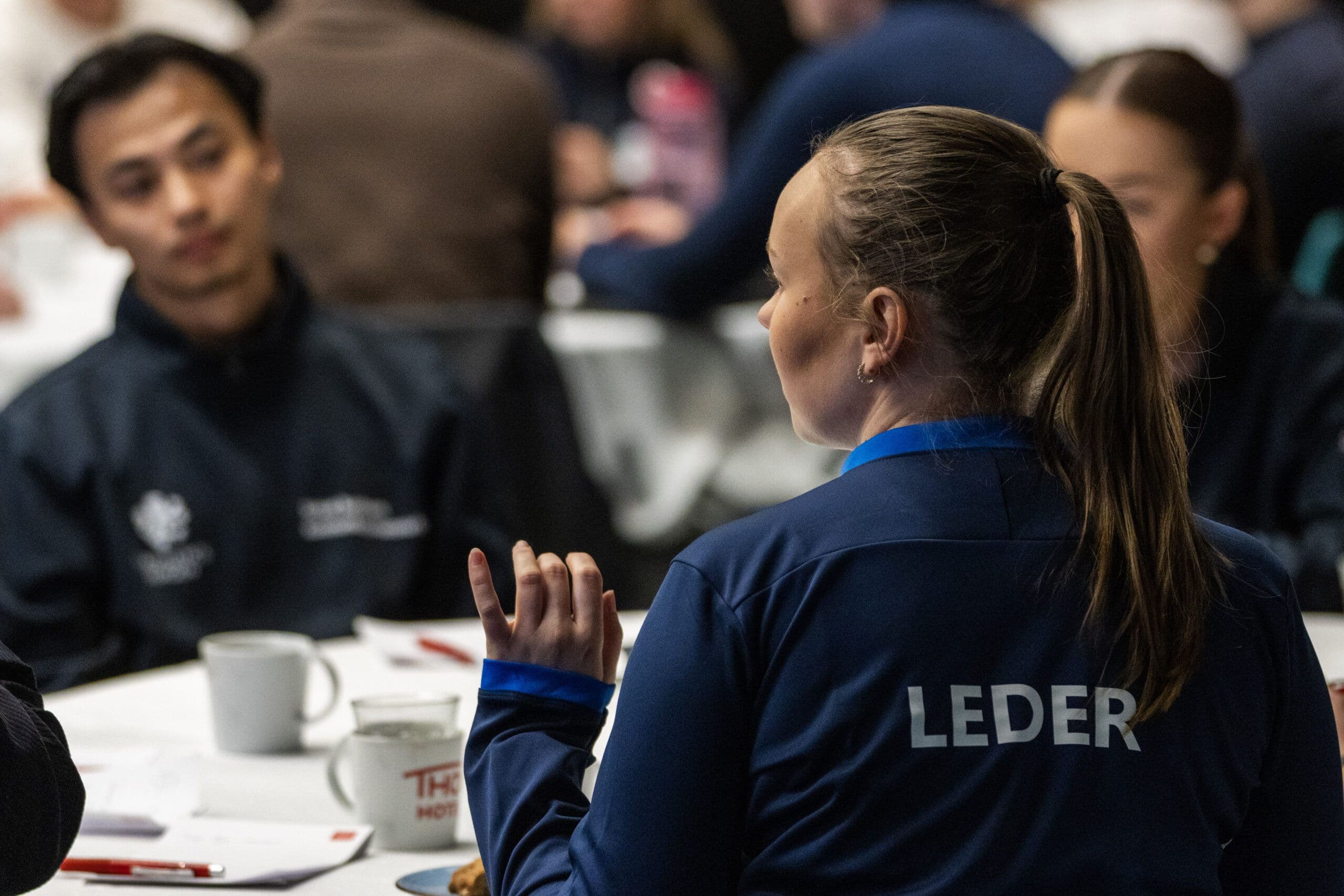 En kvinne med hestehale sett bakfra i en mørkeblå jakke med teksten «Leder», i samtale ved et bord under en samling.