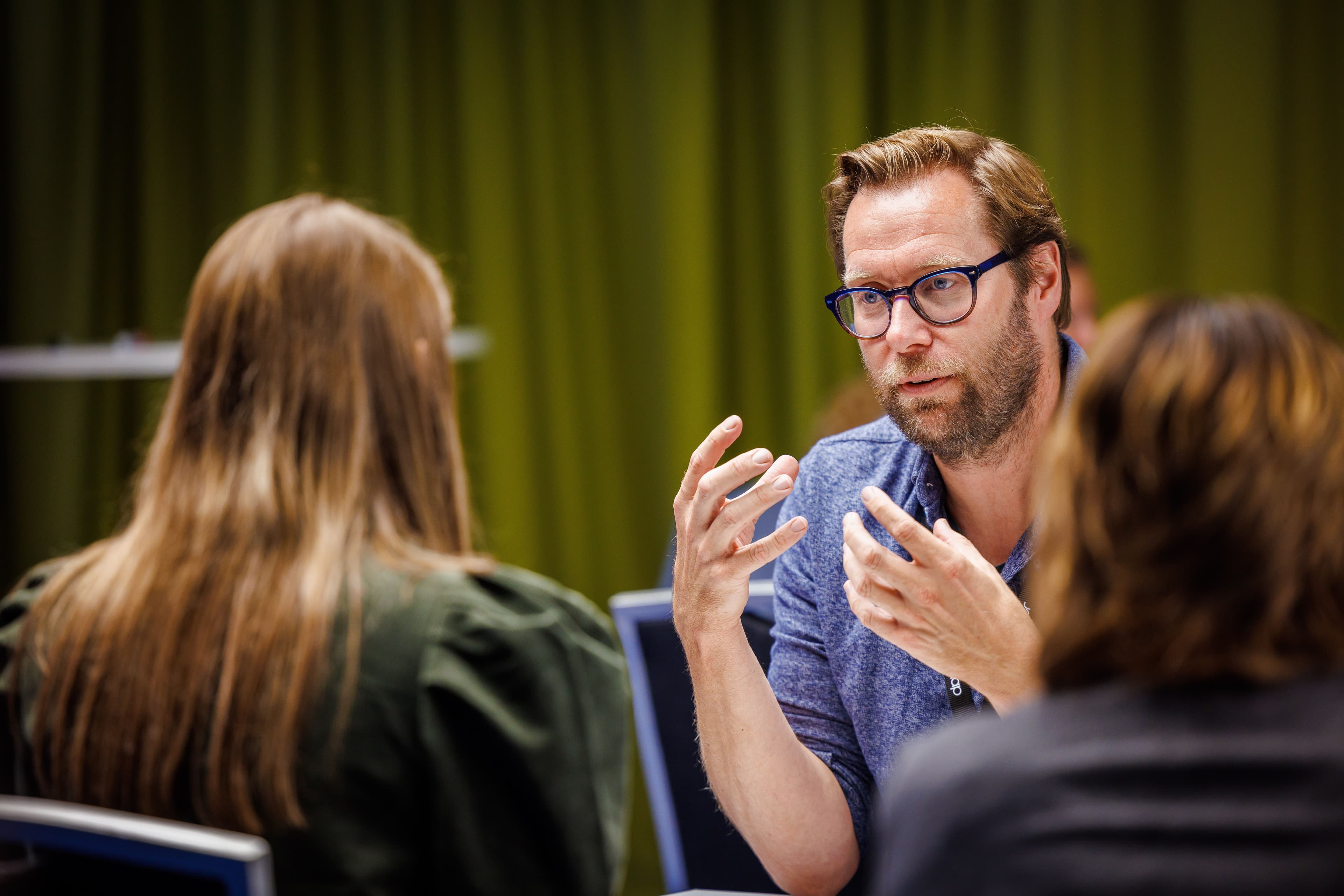 En mann med briller gestikulerer og snakker med to personer ved et bord under en samling.