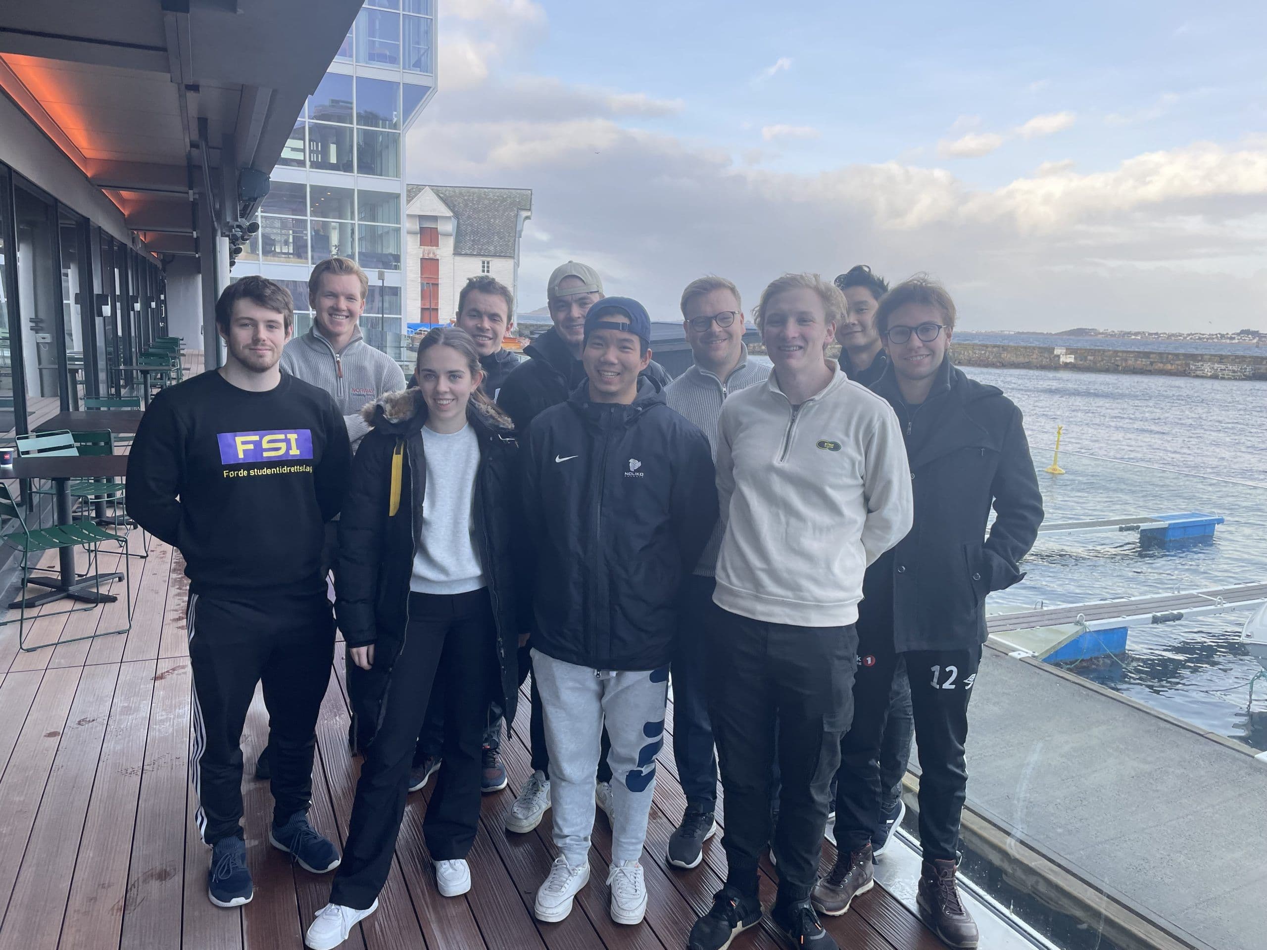 En gruppe studenter står samlet på en brygge ved sjøen med bygninger i bakgrunnen.