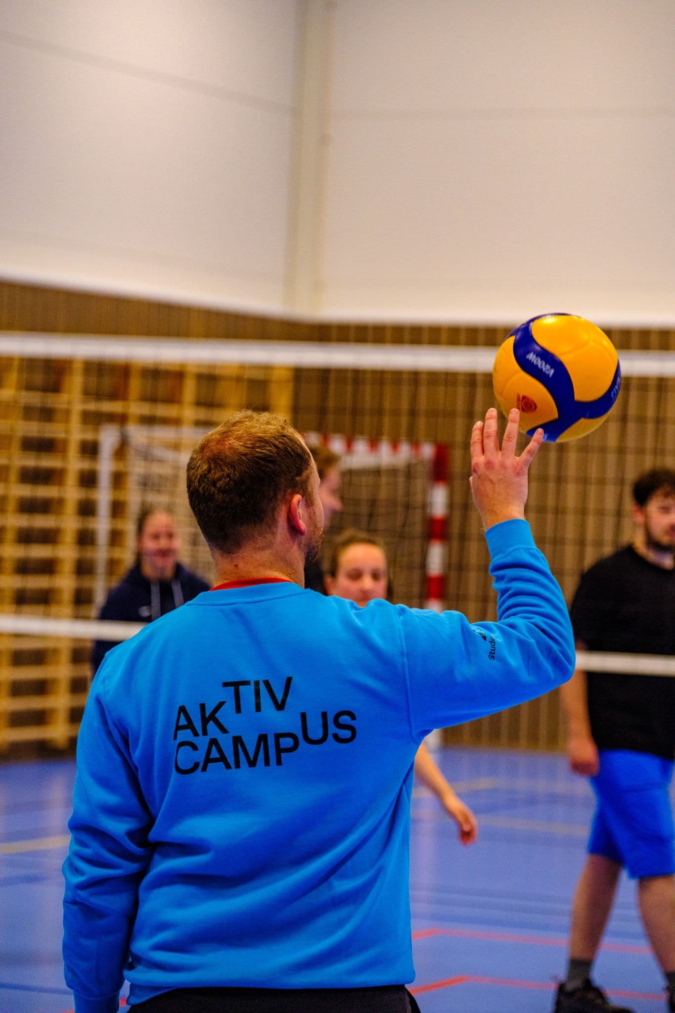En mann i blå «Aktiv Campus»-genser sett bakfra idet han skal serve en volleyball over nettet i en idrettshall.