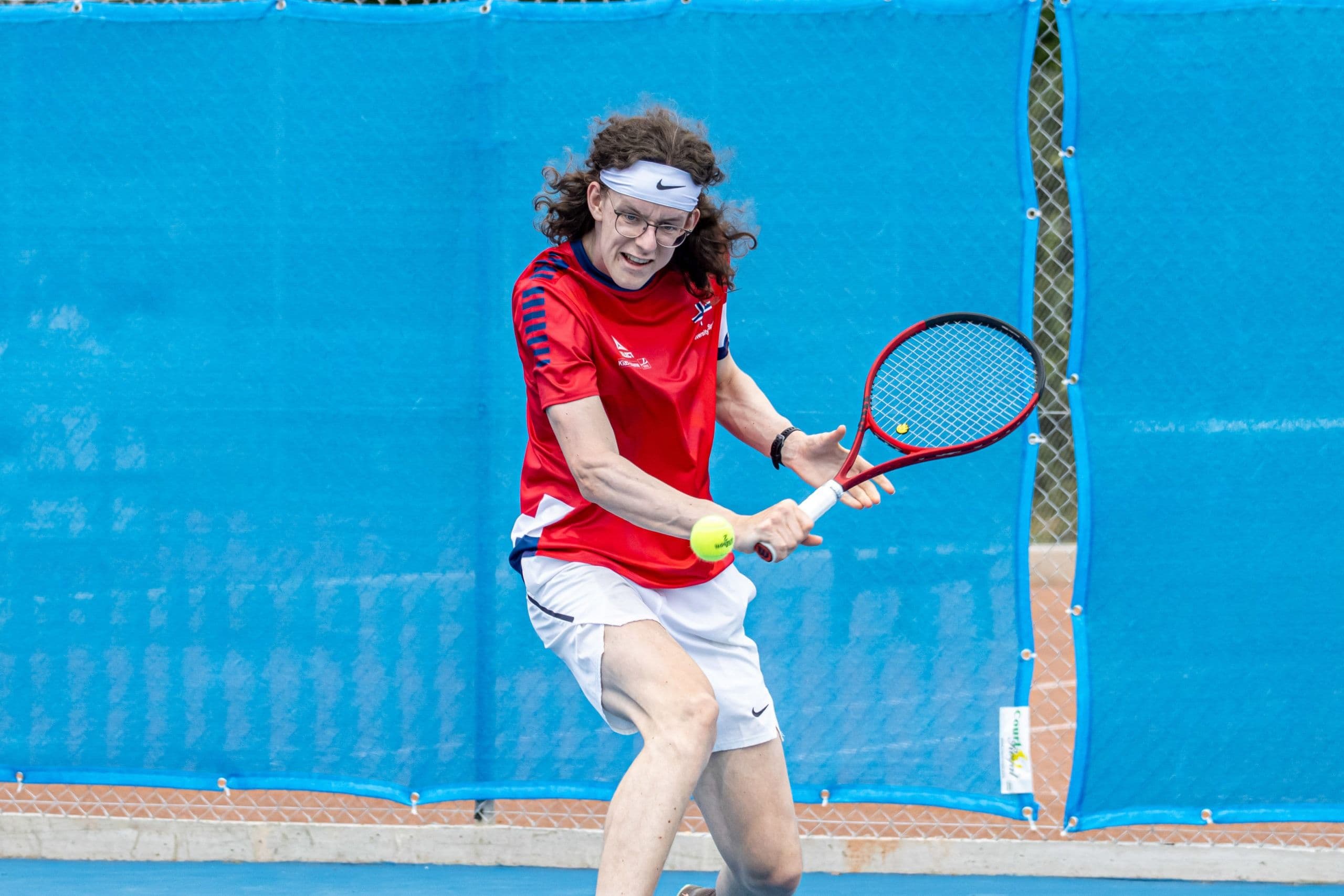 En tennisspiller i rød drakt slår en backhand på en bane med blå bakvegg.