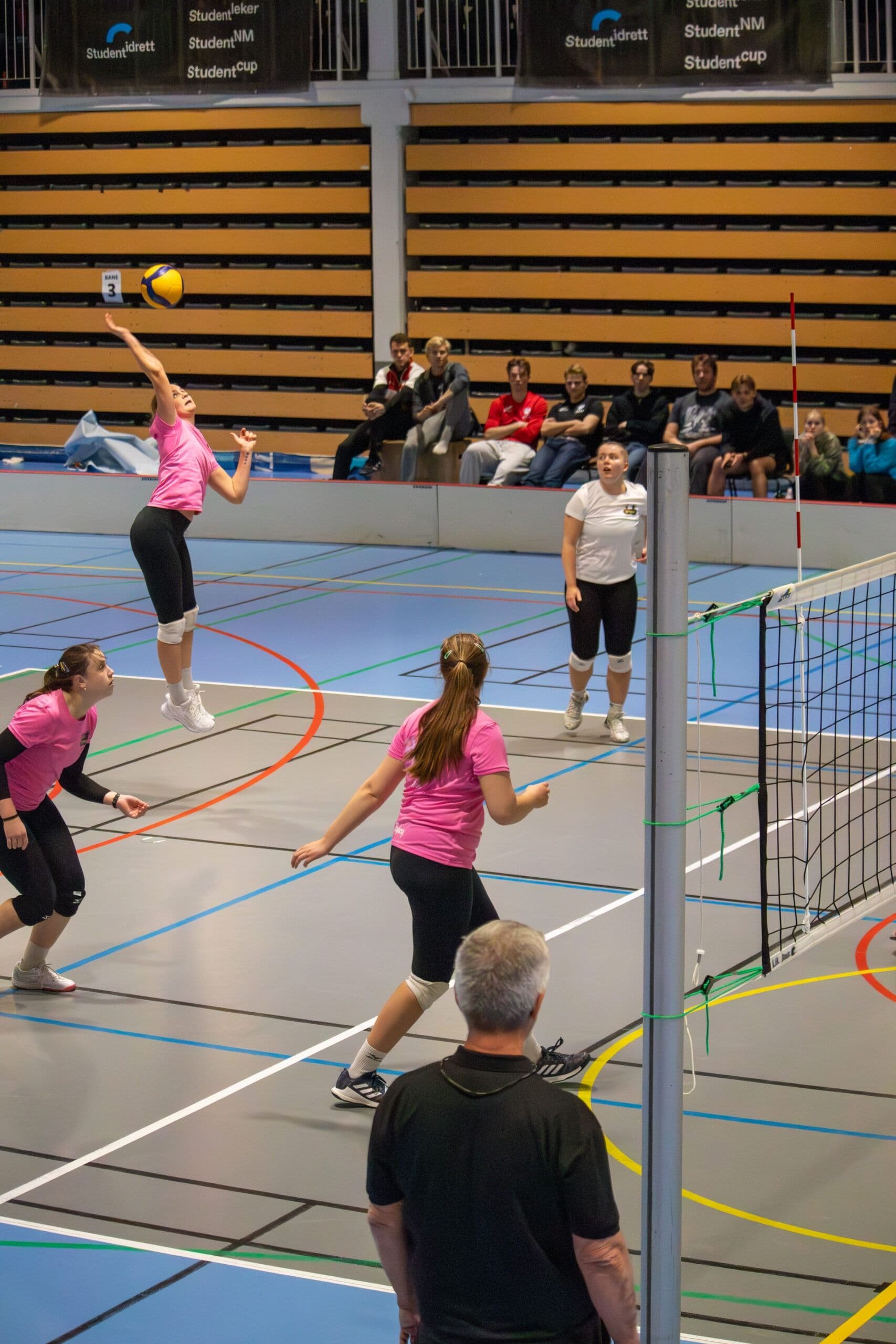 En volleyballkamp i en idrettshall der en spiller i rosa hopper for å slå ballen over nettet.
