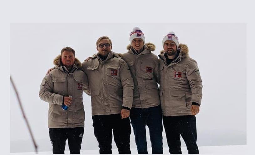 Fire menn i like beige jakker med norsk flagg står tett sammen på en snødekt fjelltopp.