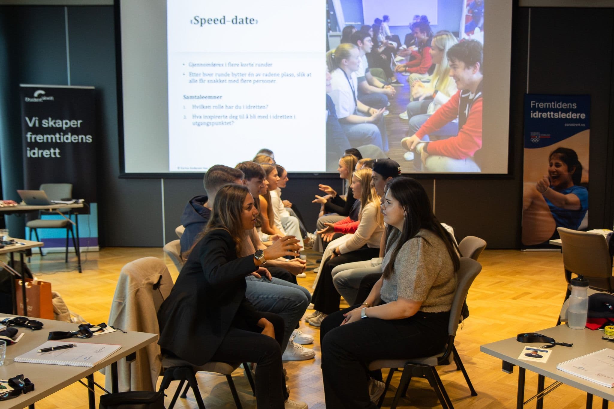 En gruppe studenter sitter i ring og samtaler under en speed-date-økt med storskjerm i bakgrunnen.