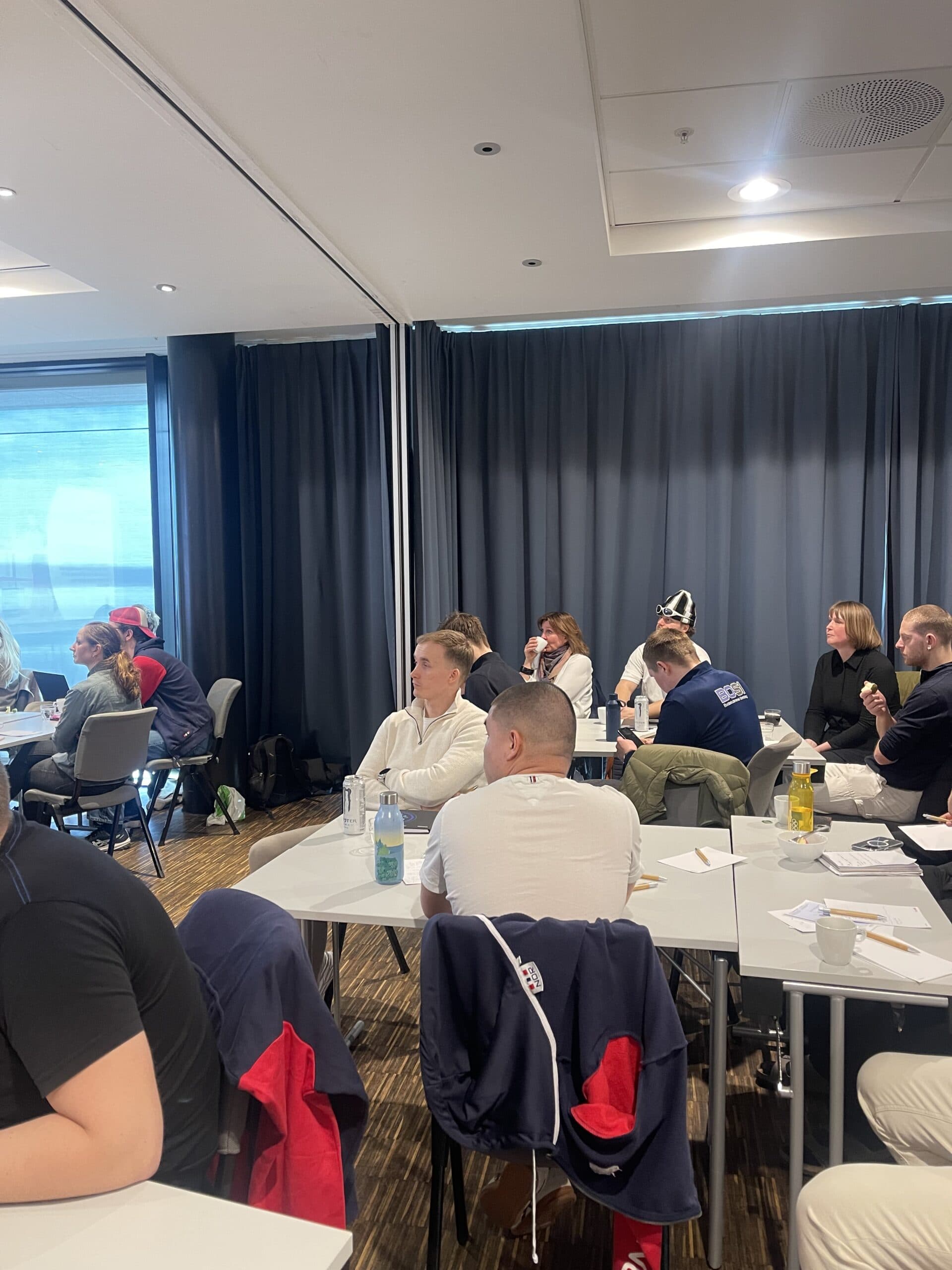 En gruppe mennesker sitter ved bord i et møterom og følger med på en presentasjon.