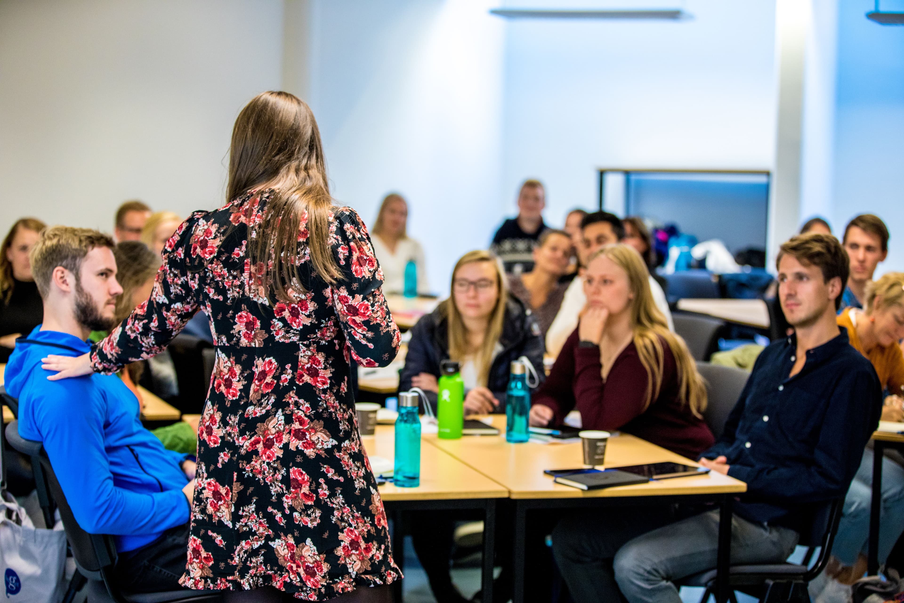 En kvinne holder et innlegg foran en gruppe studenter som sitter ved bord i et klasserom.