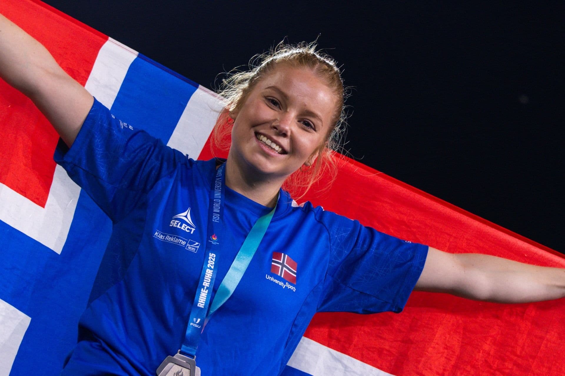 En smilende utøver med medalje holder et norsk flagg utstrakt bak seg.