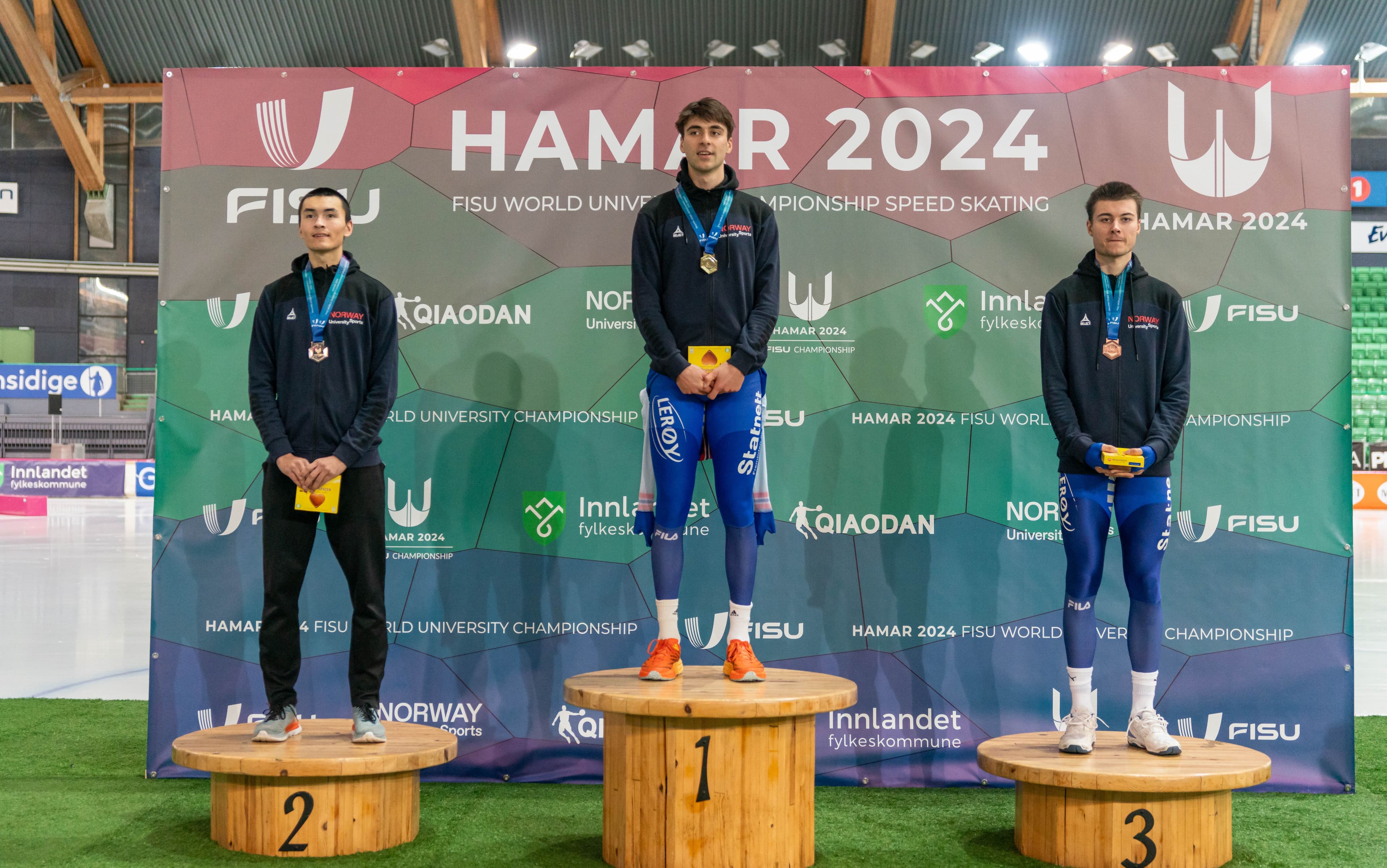 Tre skøyteløpere med medaljer står på seierspallen foran en vegg med teksten Hamar 2024 FISU World University Championship Speed Skating.