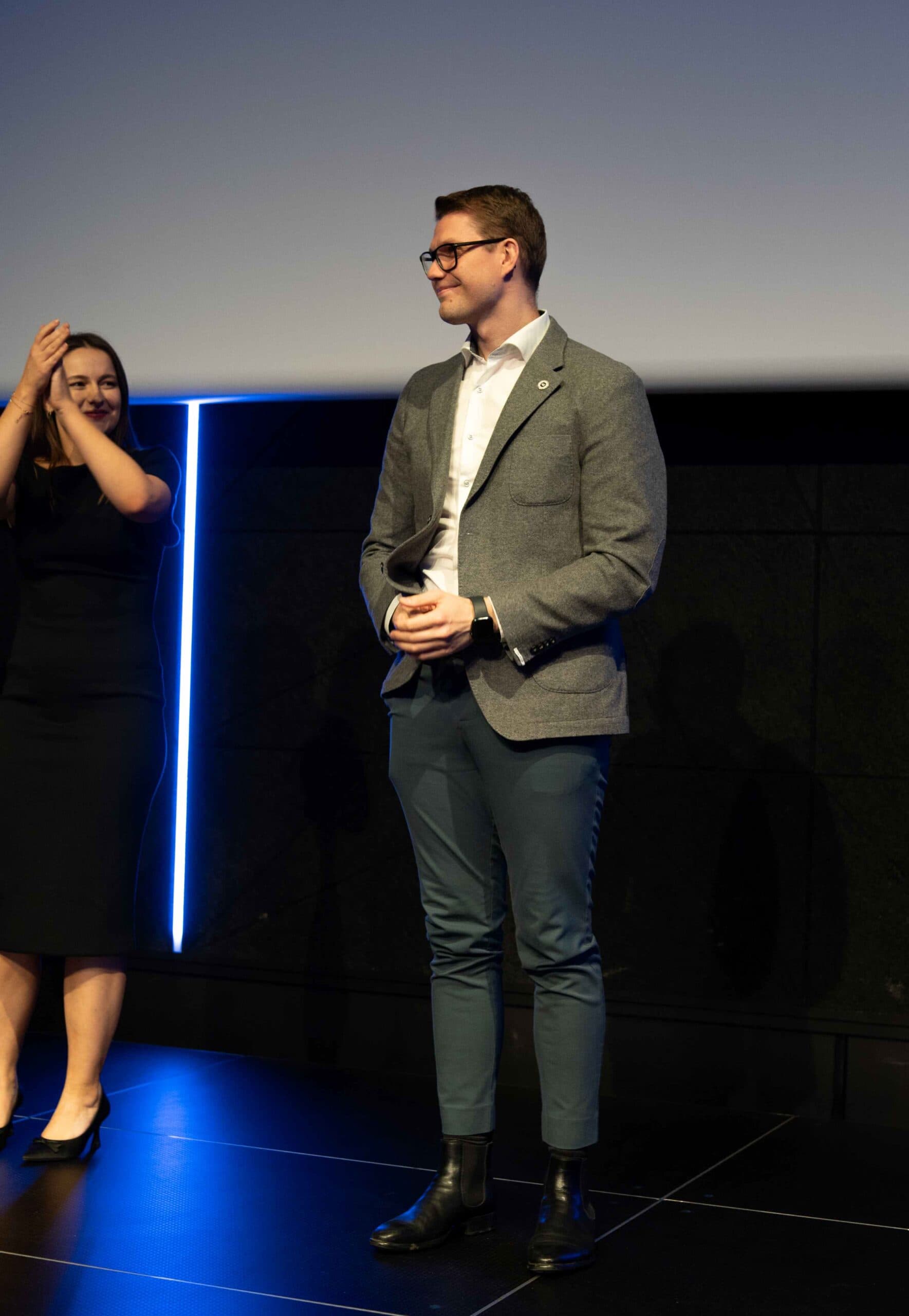 En mann i grå dressjakke står på en scene mens en kvinne ved siden av applauderer.