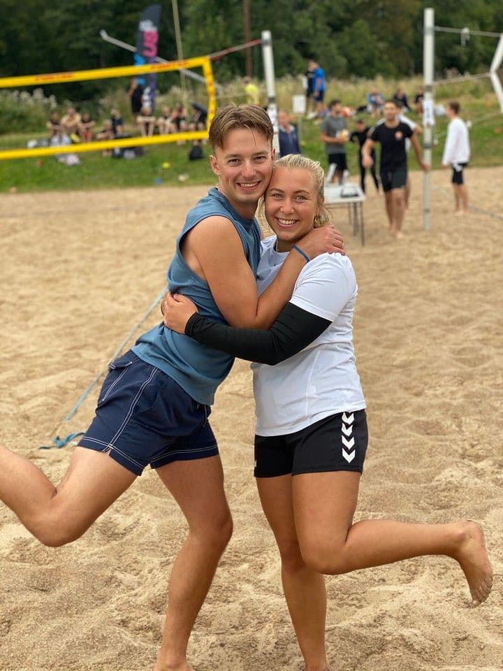 En mann og en kvinne omfavner hverandre og smiler på en sandvolleyballbane.
