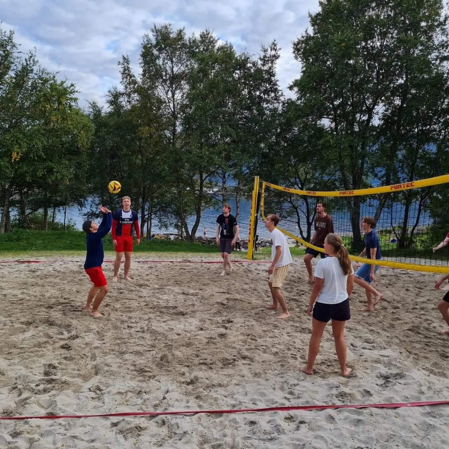 En gruppe studenter spiller sandvolleyball på en strand ved vannet.