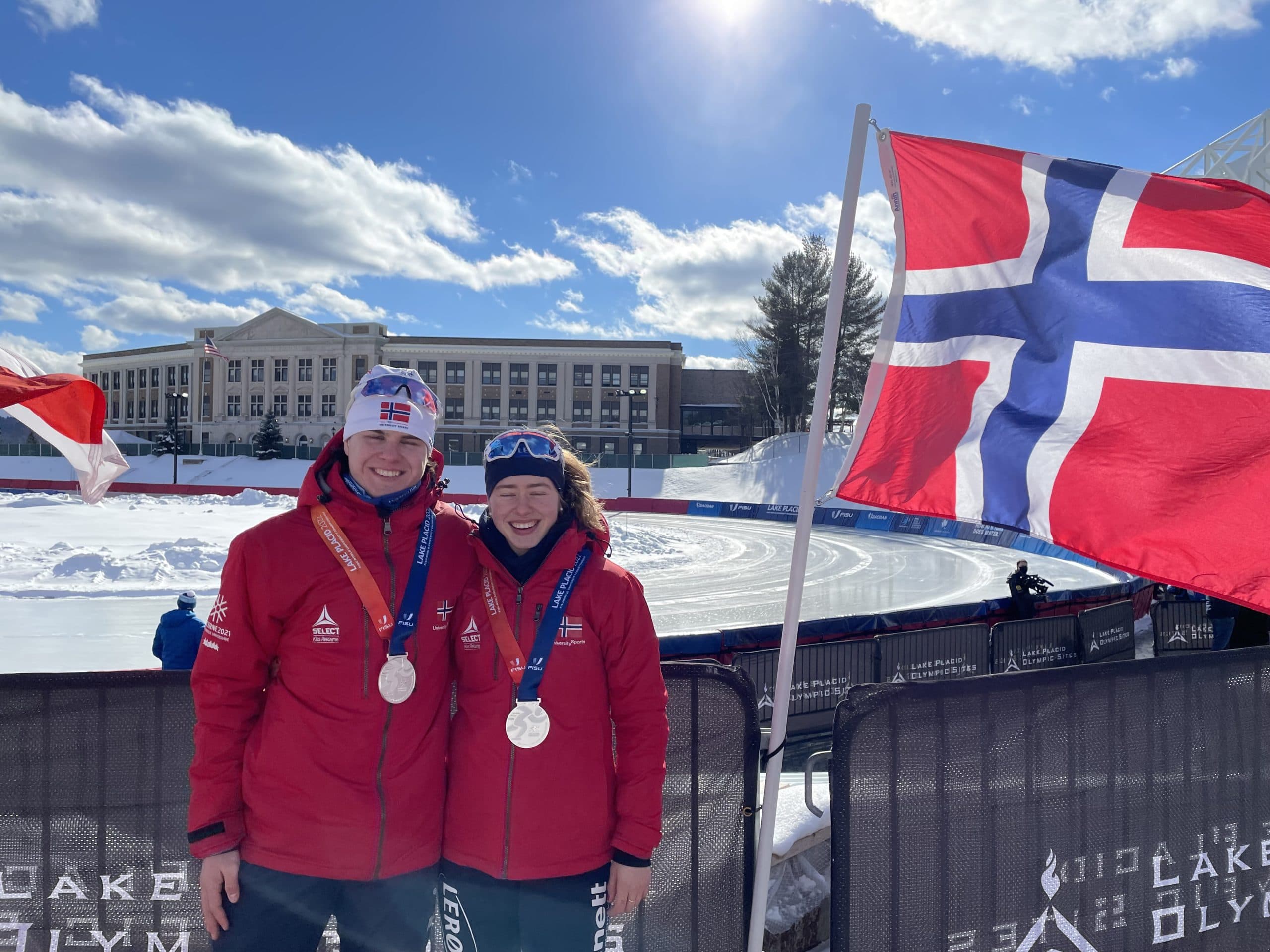 To utøvere i røde jakker viser fram medaljene sine foran et norsk flagg utendørs om vinteren.