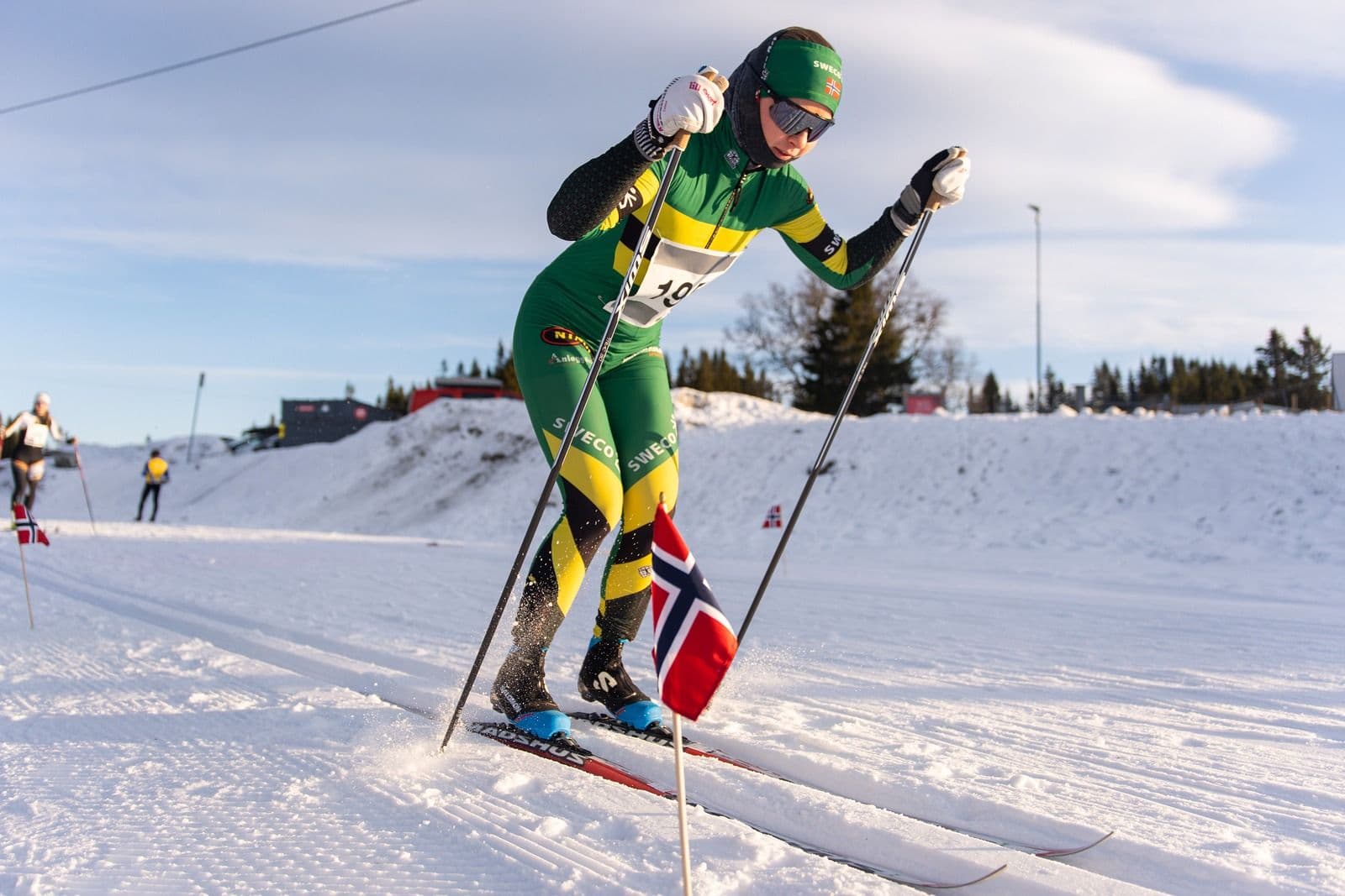 En langrennsløper i grønn og gul drakt staker seg fram i løypa, med et norsk flagg ved sporet.