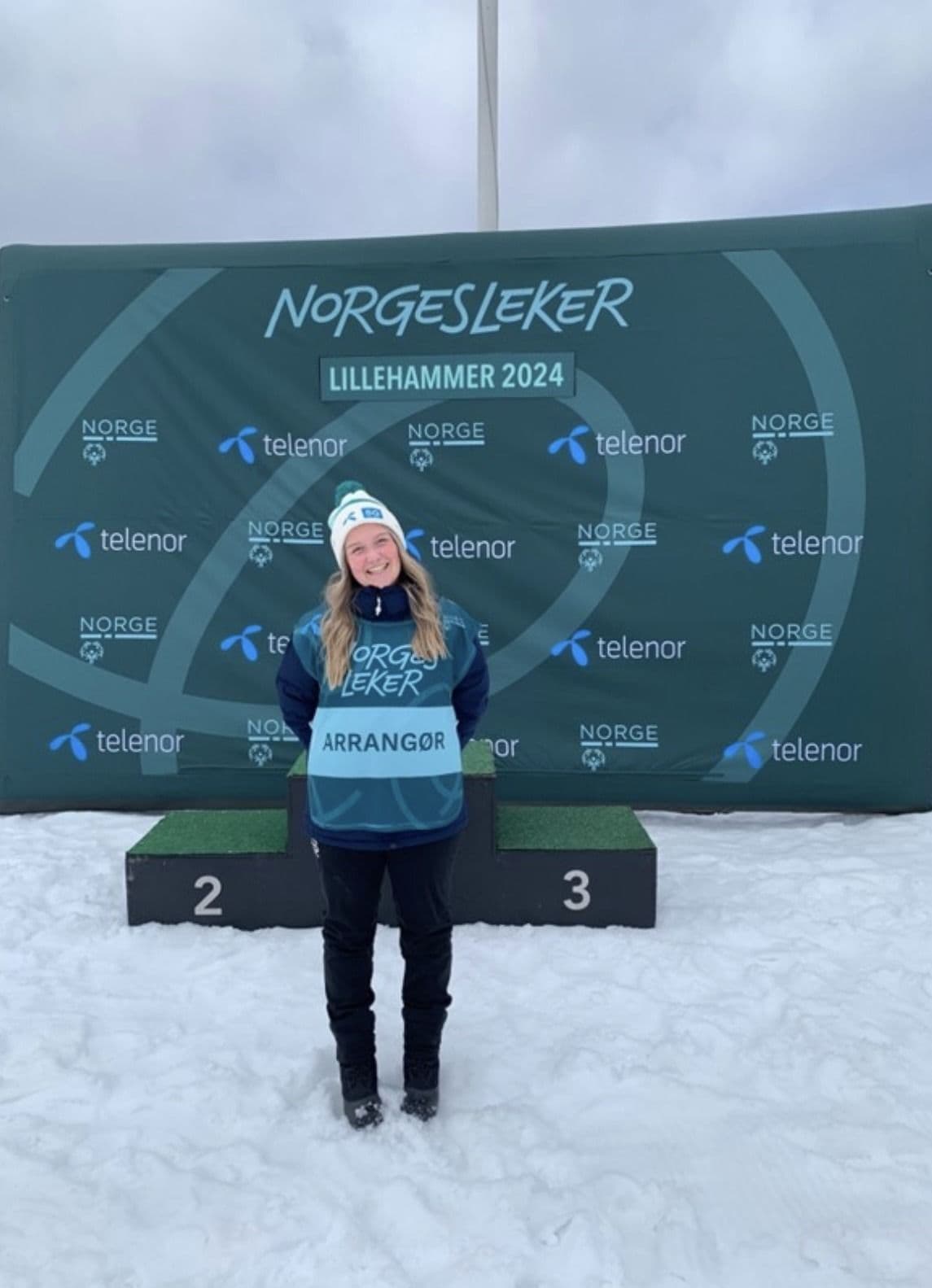 En kvinne med arrangørvest står foran en vegg med logoen til Norgesleker Lillehammer 2024.