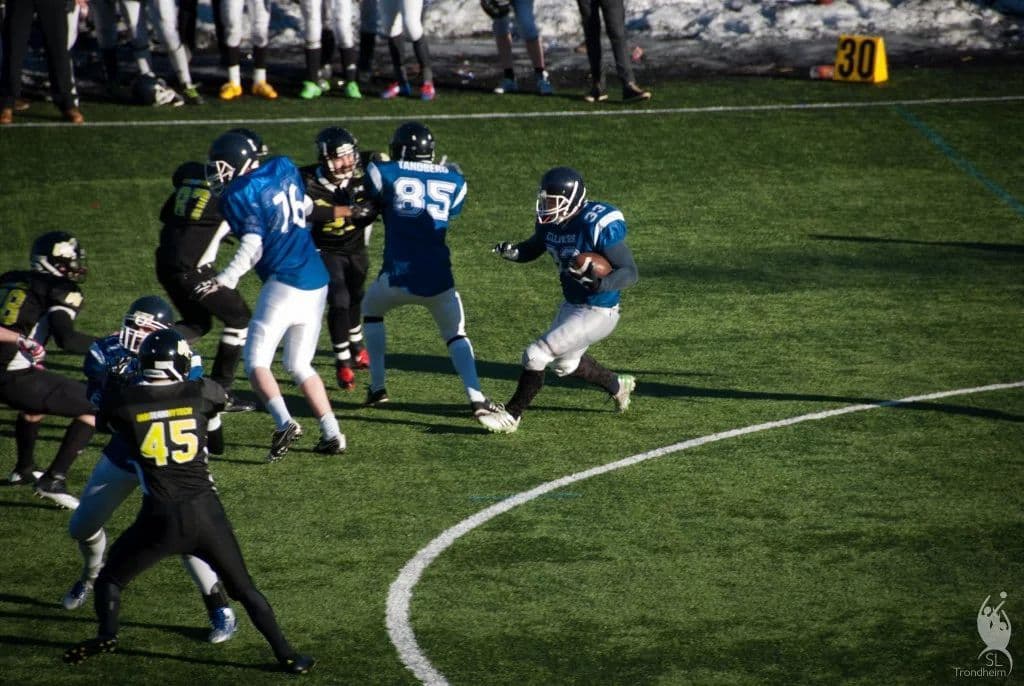 Spillere i amerikansk fotball med hjelm og skulderbeskyttere i dueller under en kamp på kunstgress.