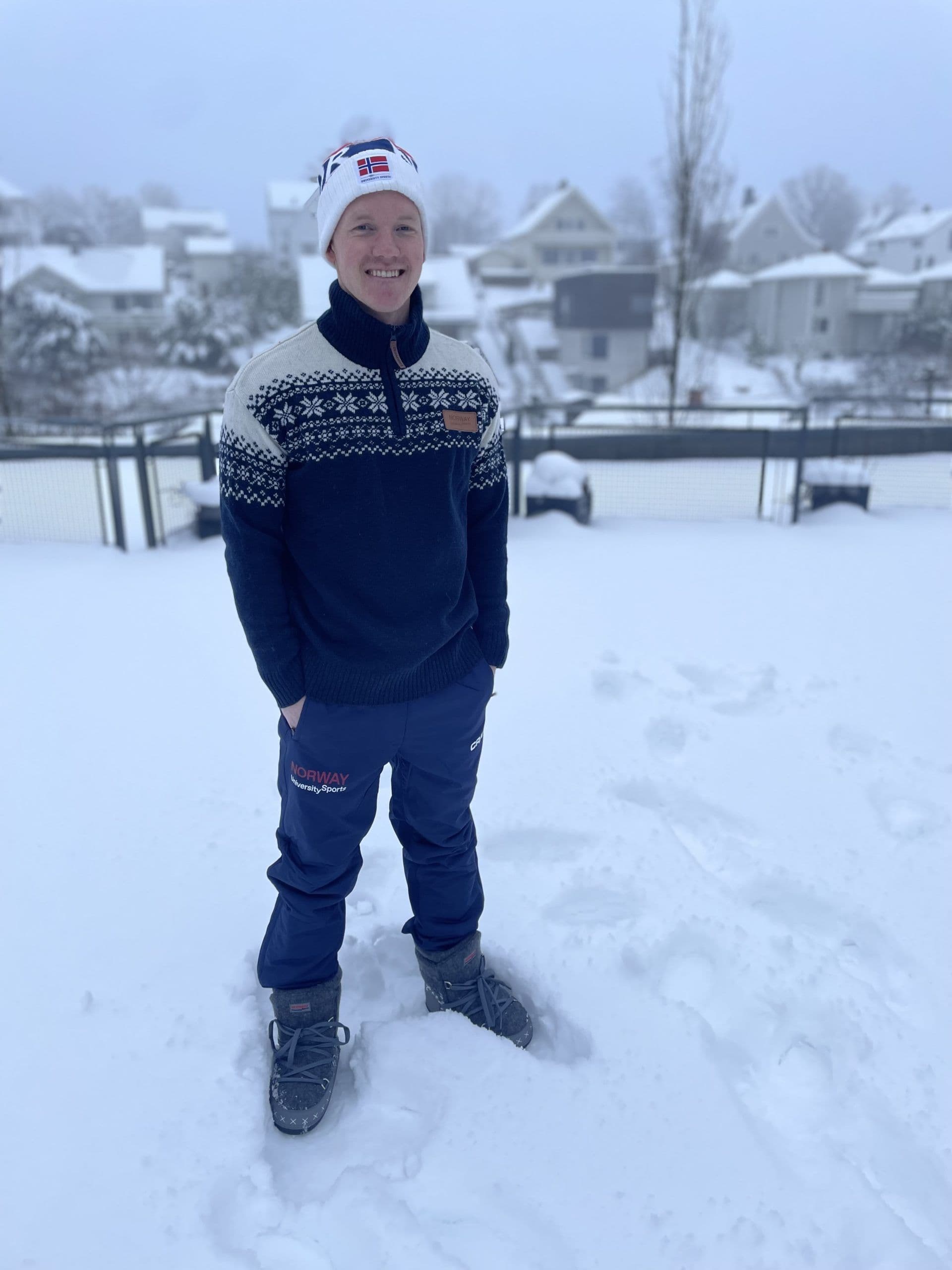 En smilende mann i strikkegenser og lue med norsk flagg står i snøen i et boligstrøk.