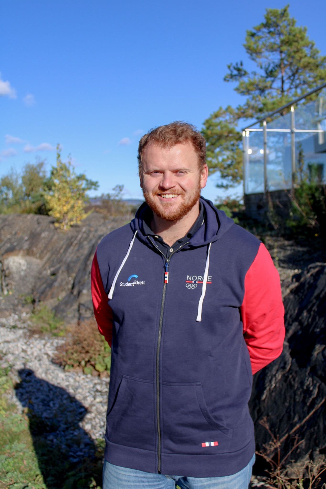 Portrett av en smilende mann med skjegg i Studentidrett- og Norge-jakke utendørs i sola.