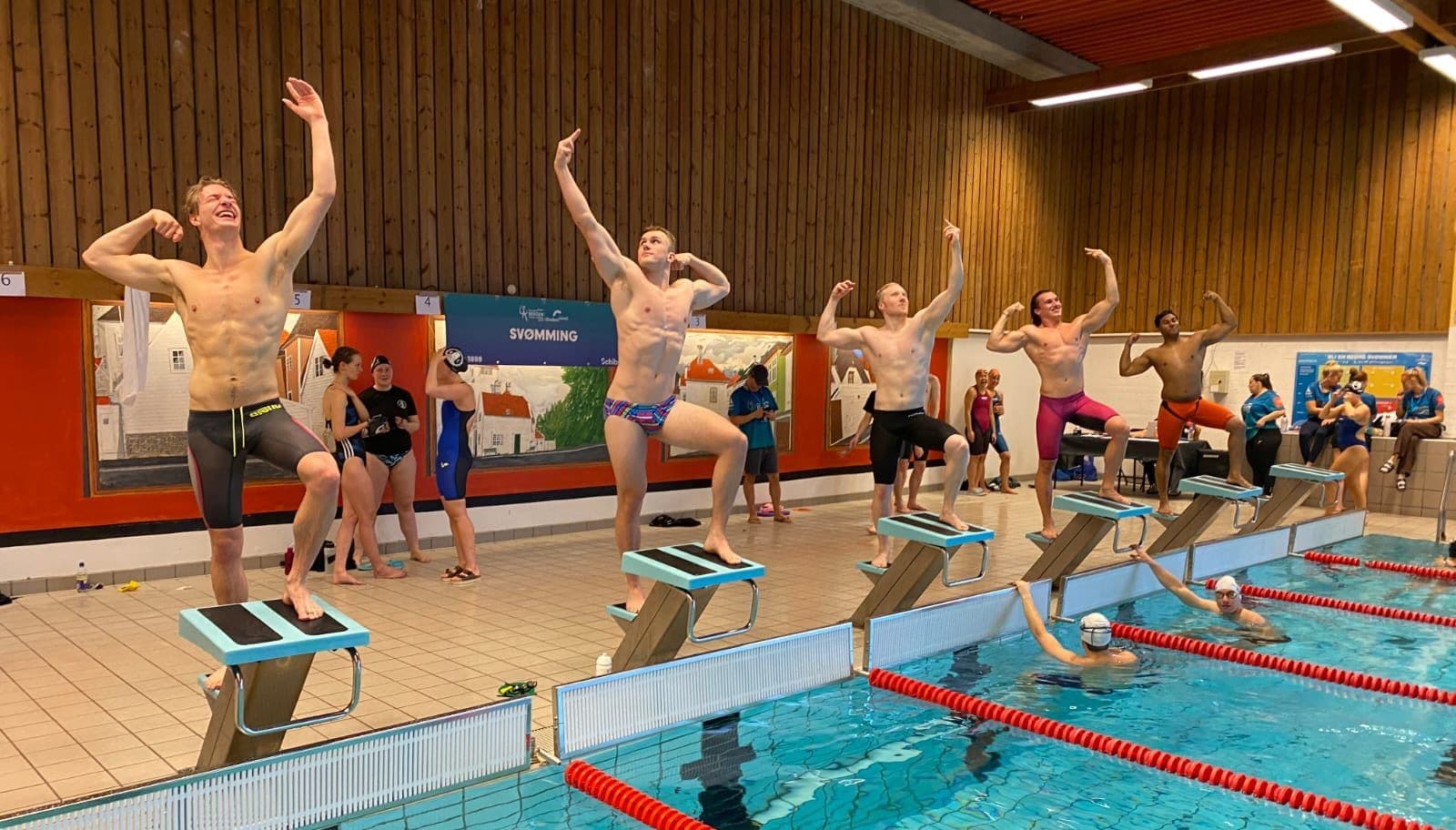 Flere svømmere står på startpallene ved bassenget og poserer med spente muskler.
