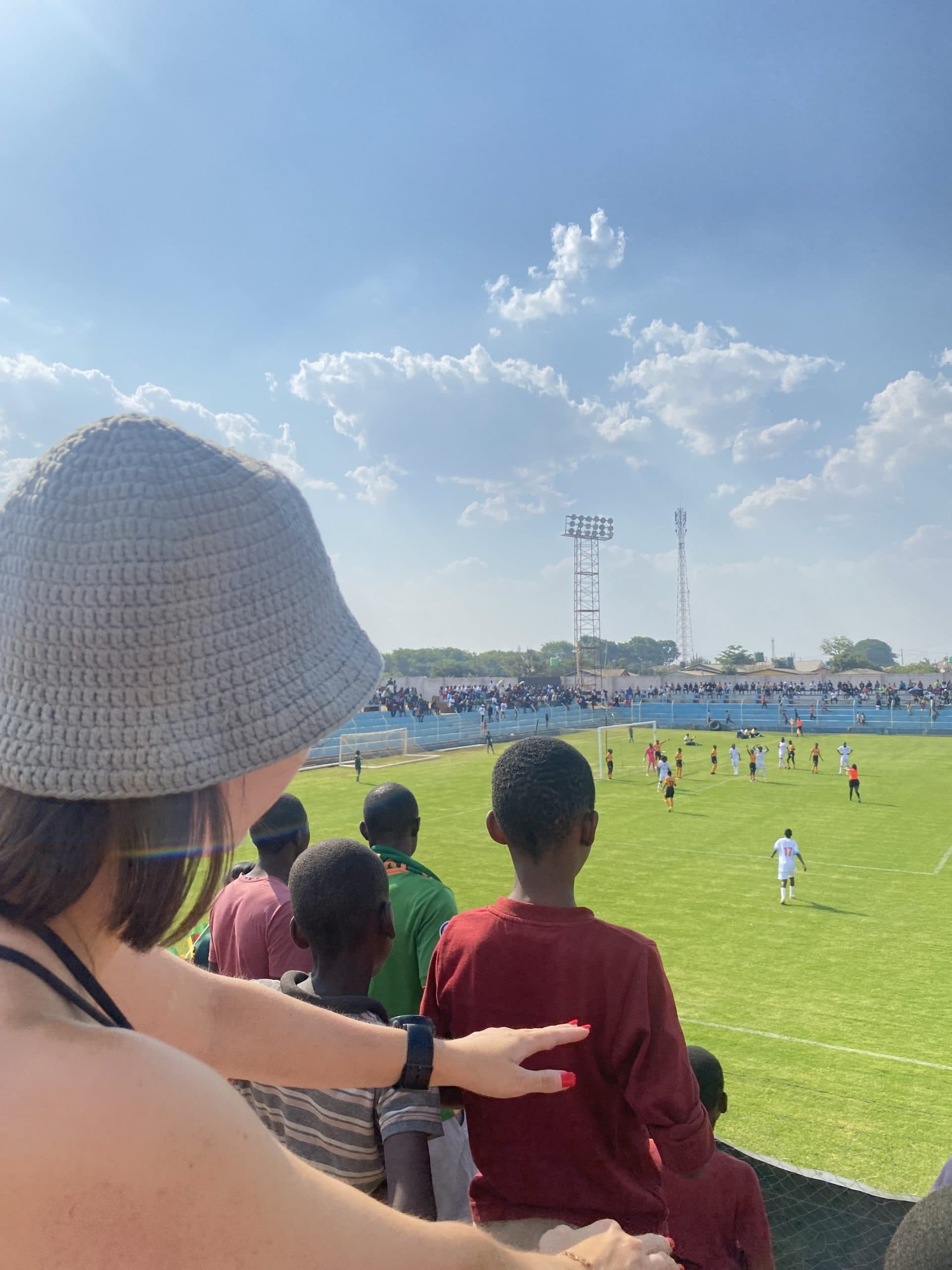 En person med solhatt følger en fotballkamp fra tribunen på et stadion i sterkt solskinn.