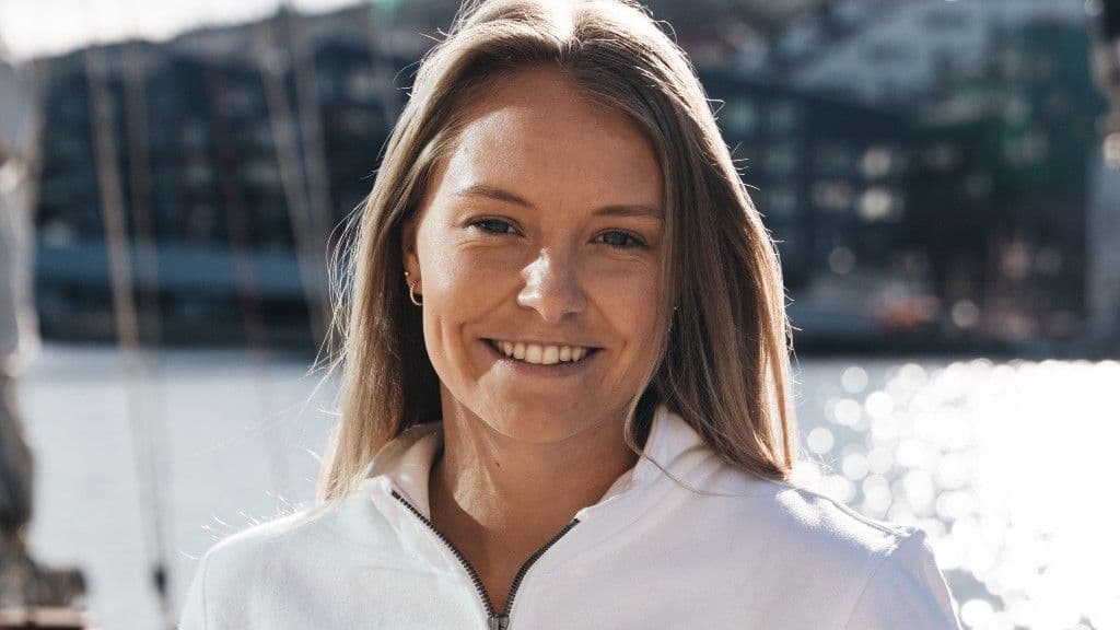 Portrett av en smilende kvinne i hvit jakke med en havn i bakgrunnen.