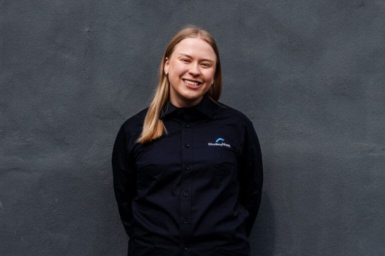 Portrett av en smilende kvinne i svart skjorte med Studentidrett-logo mot en mørk vegg.