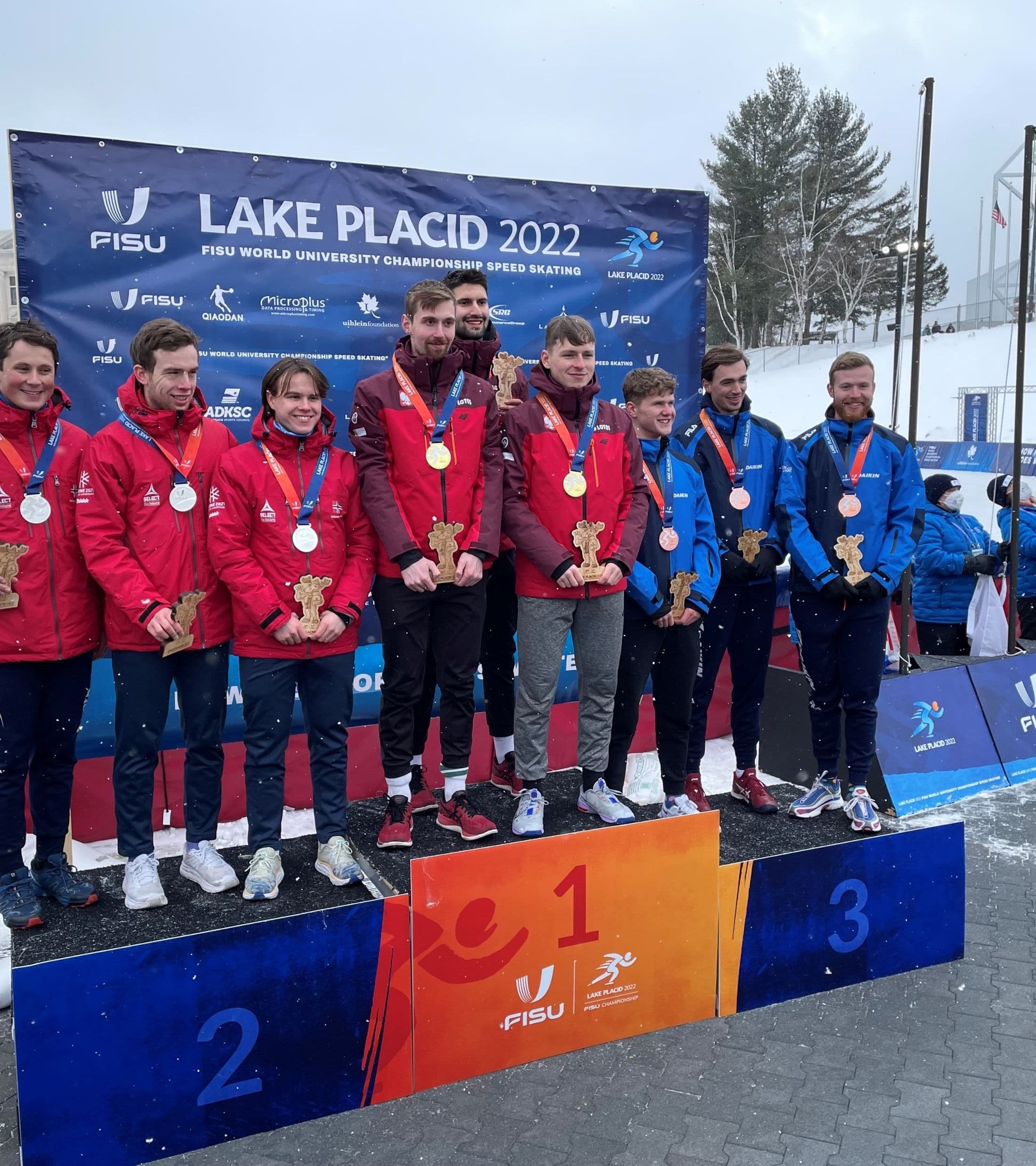 En gruppe skøyteløpere med medaljer på seierspallen foran en FISU-vegg med teksten Lake Placid 2022.