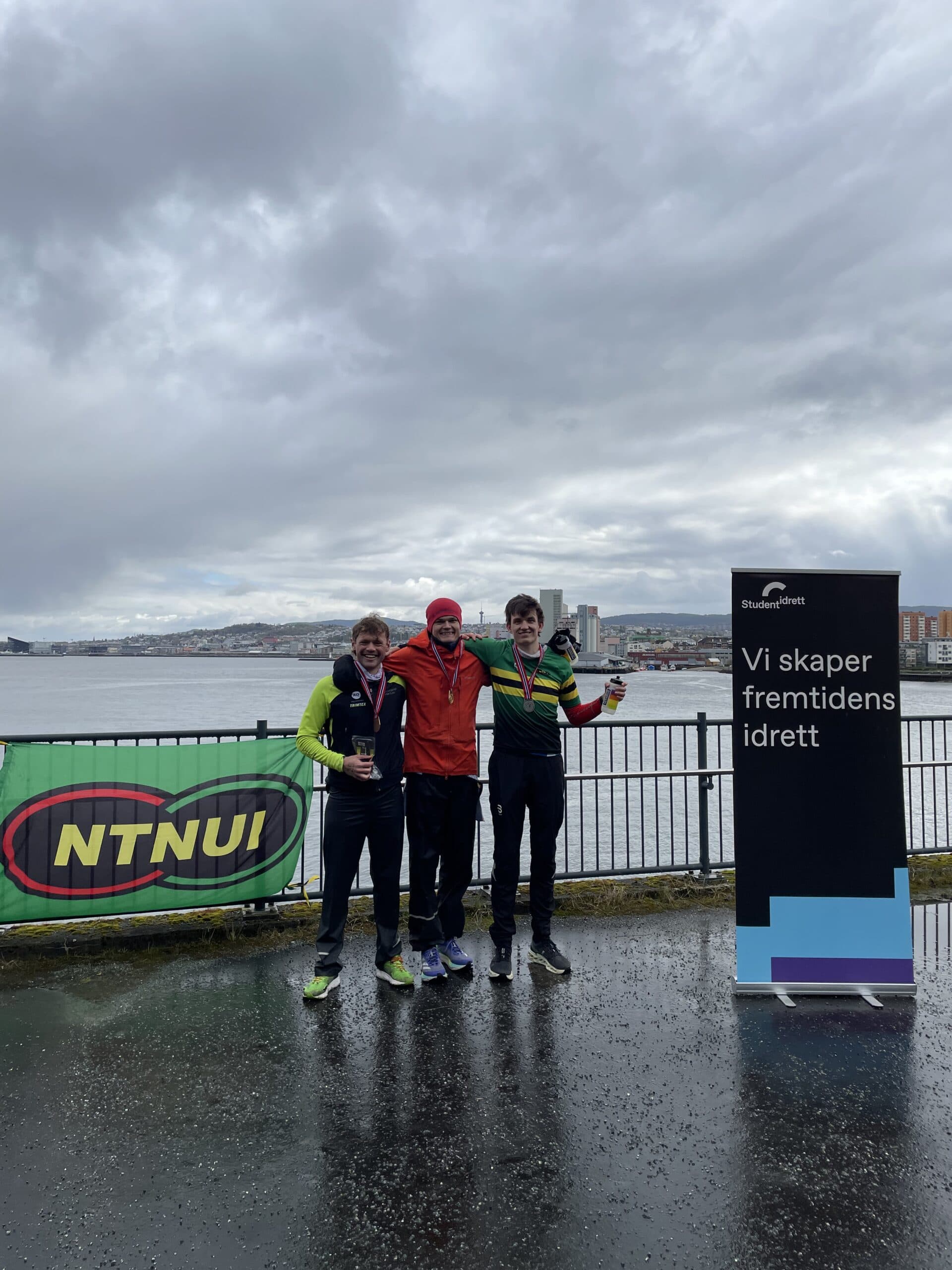 Tre løpere med medaljer poserer ved sjøkanten foran en NTNUI-banner og en Studentidrett-rollup.