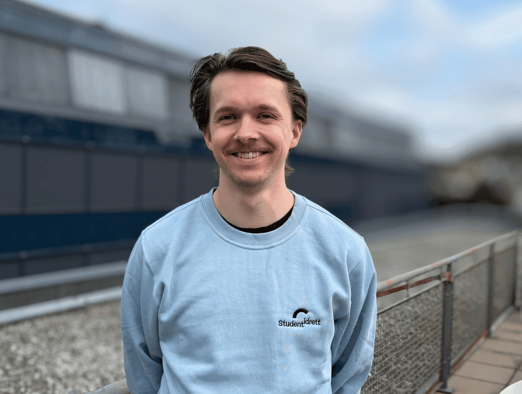 Portrett av en smilende mann i lyseblå genser med Studentidrett-logo, utendørs.
