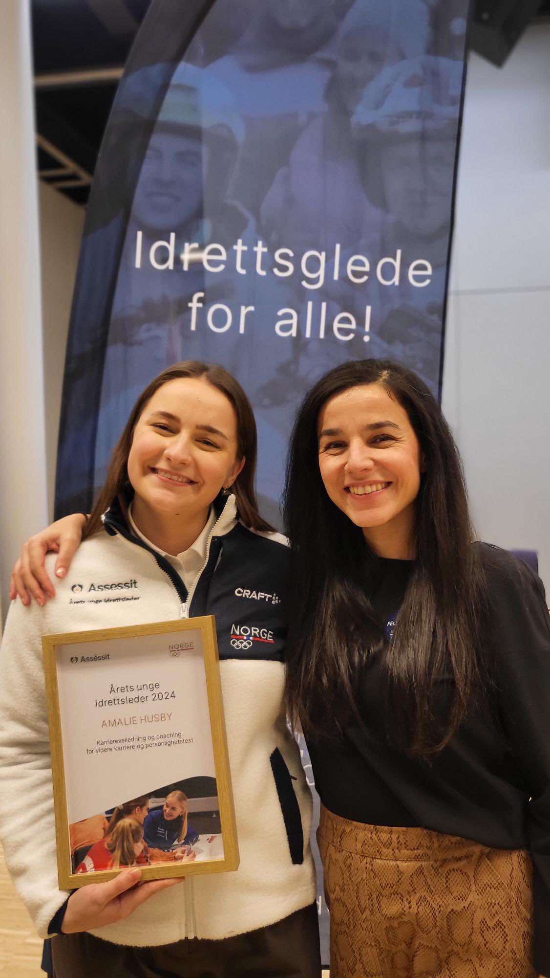 To smilende kvinner poserer med et diplom for Årets unge idrettsleder 2024 foran en banner med teksten Idrettsglede for alle.