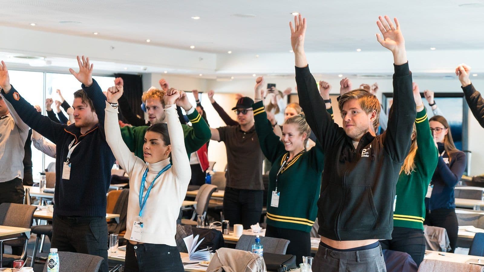 En gruppe studenter reiser seg med armene i været under en samling i et møterom.