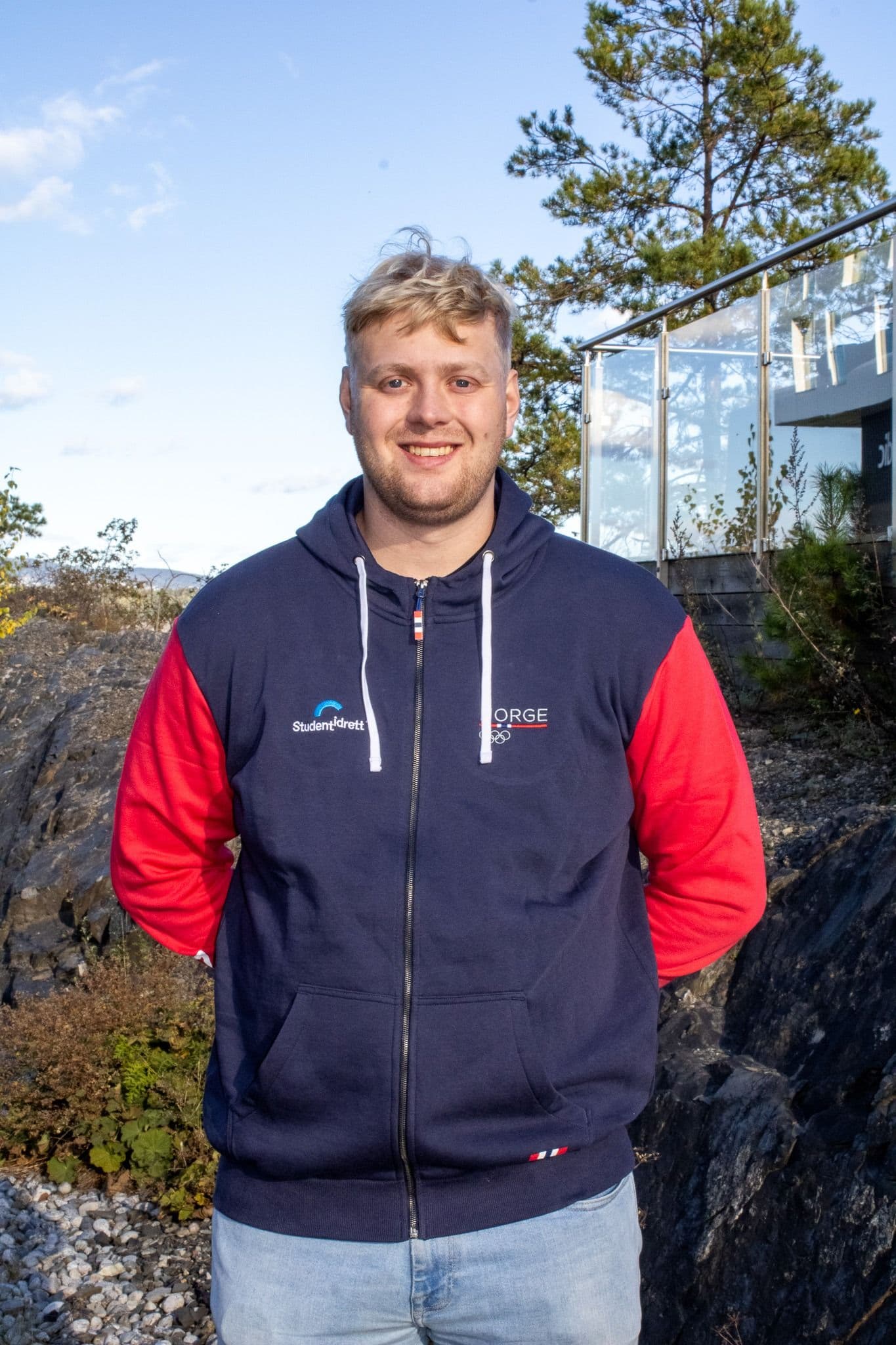 Portrett av en smilende mann i mørk jakke med Studentidrett- og Norge-logo, utendørs.