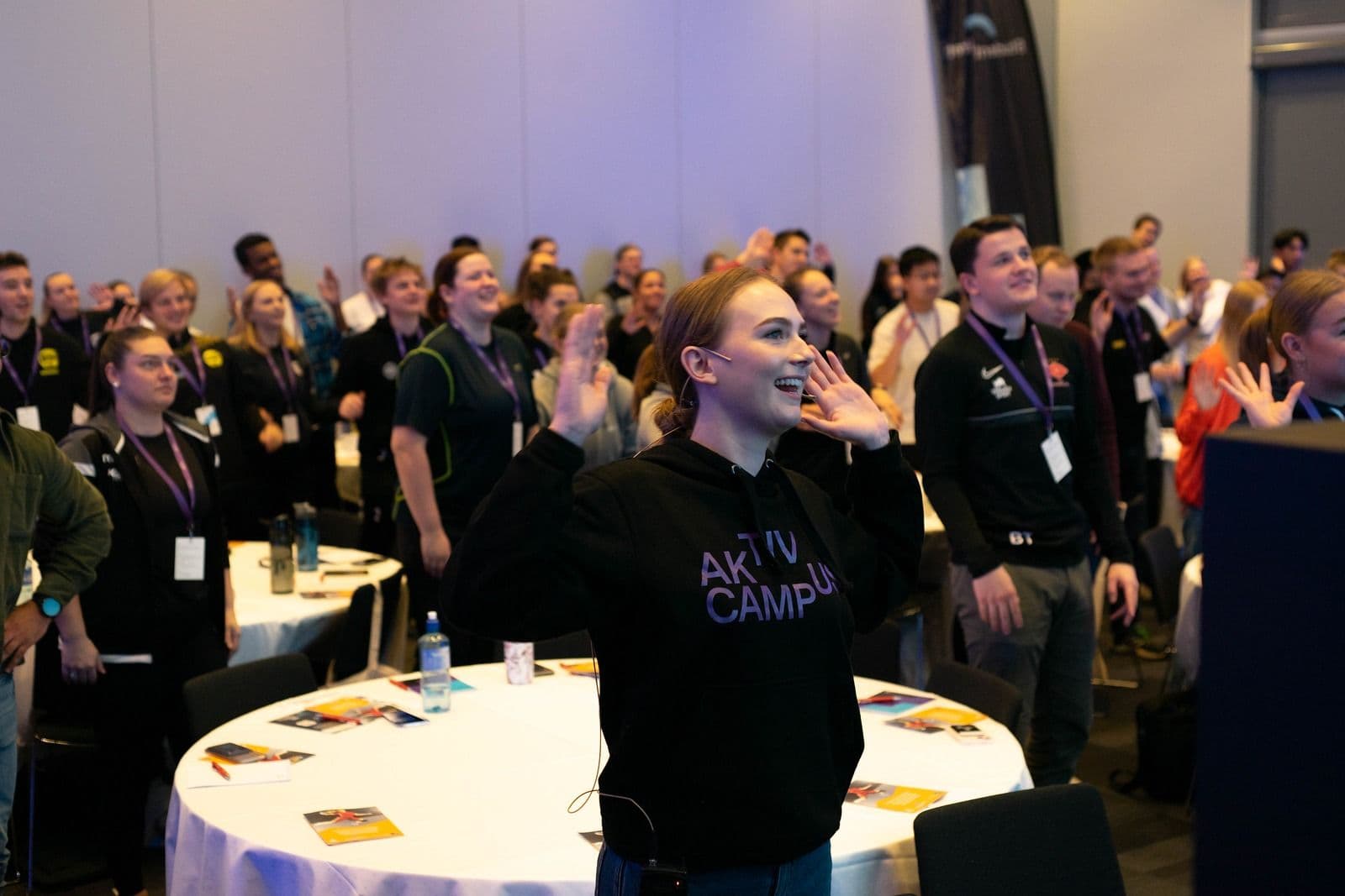 En kvinne i Aktiv Campus-genser jubler foran en stor forsamling i en konferansesal.