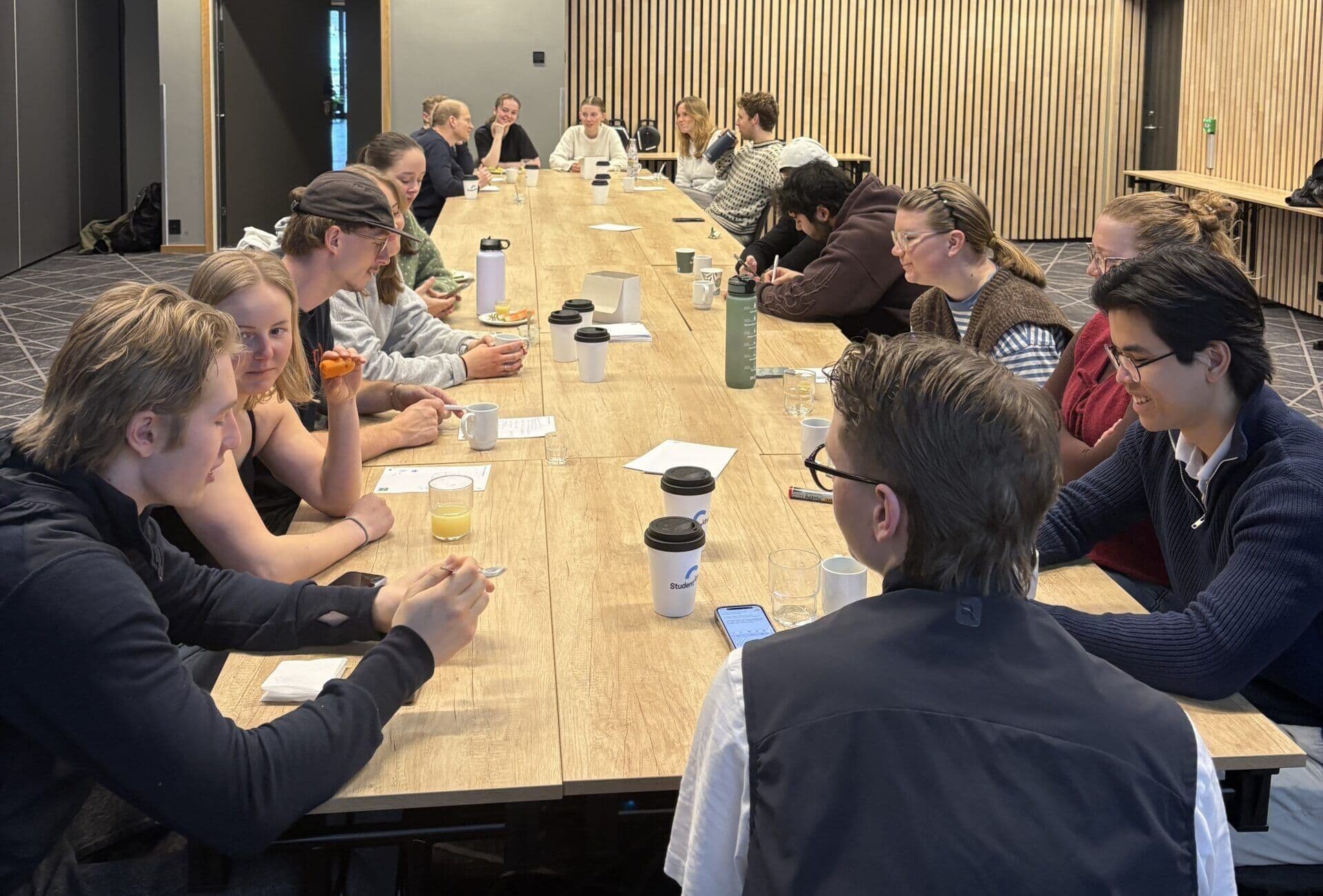 En gruppe studenter sitter i møte rundt et langt bord med kaffekopper og papirer.