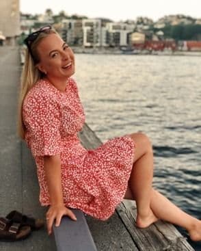 Portrett av en kvinne i rød sommerkjole som sitter smilende på en brygge ved vannet.