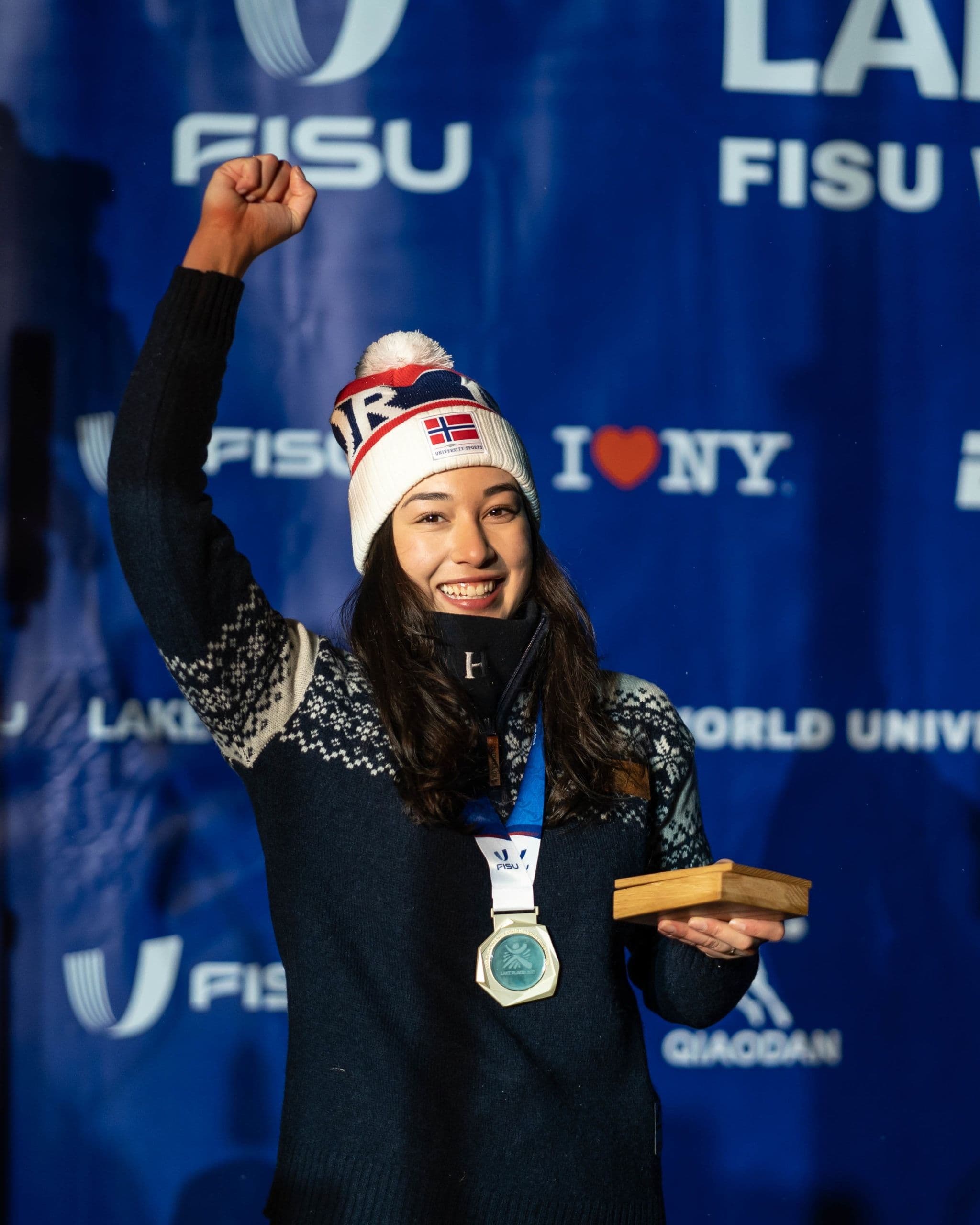 En kvinne med norsk lue løfter armen i været og holder en medalje foran en FISU-vegg fra Lake Placid.