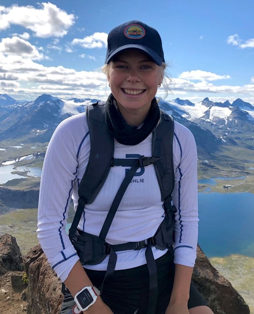 En kvinne med caps og tursekk står smilende på en fjelltopp med utsikt over fjell og vann.