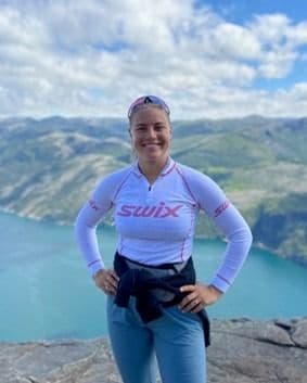 En kvinne i treningstopp står smilende på et fjell med utsikt over en fjord.