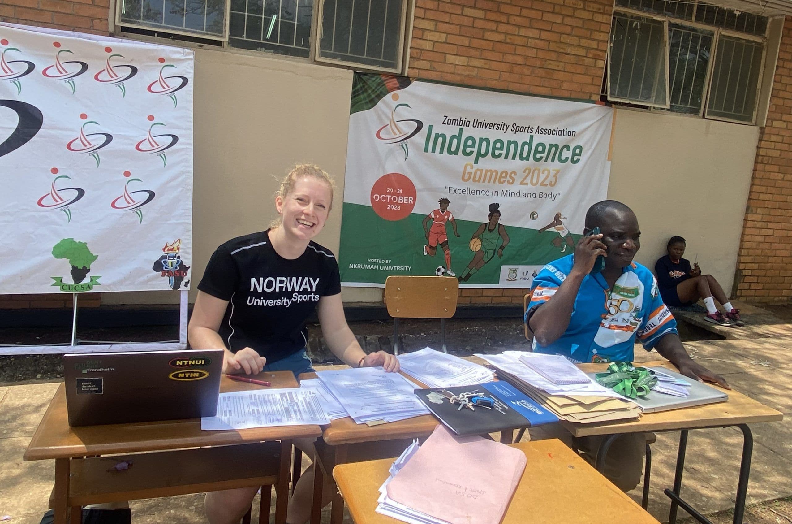 En kvinne i Norway University Sports-genser sitter smilende ved et registreringsbord under Zambia University Sports Independence Games 2023.