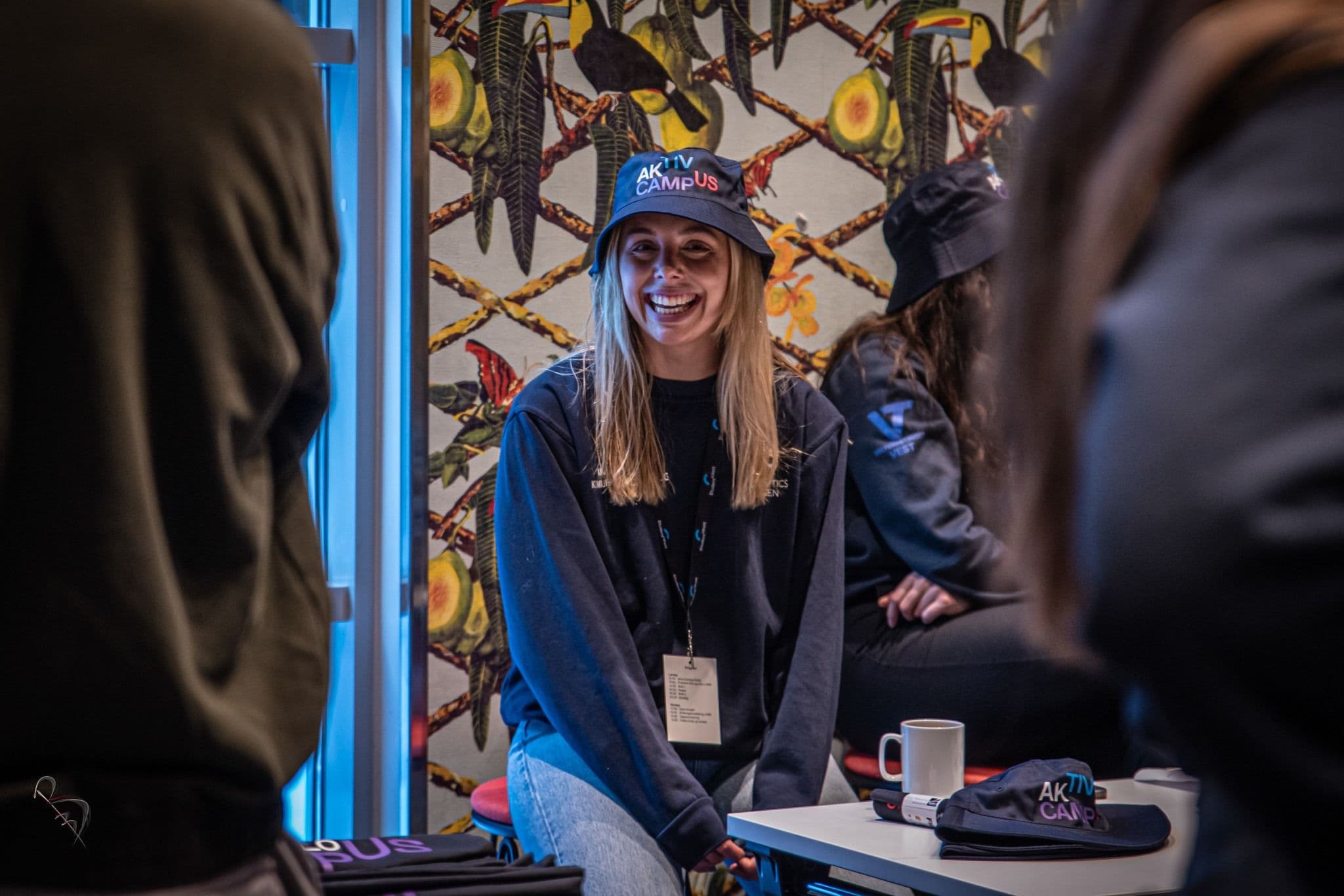 En kvinne i Aktiv Campus-caps smiler under en samtale på et arrangement.