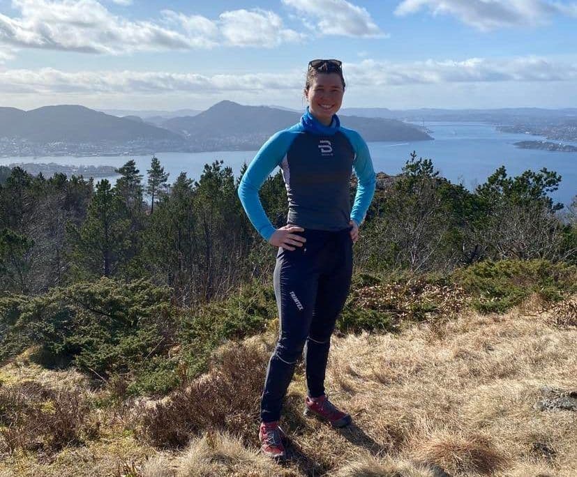 En kvinne i treningsklær står på en fjelltopp med utsikt over en fjord og skogkledde åser.