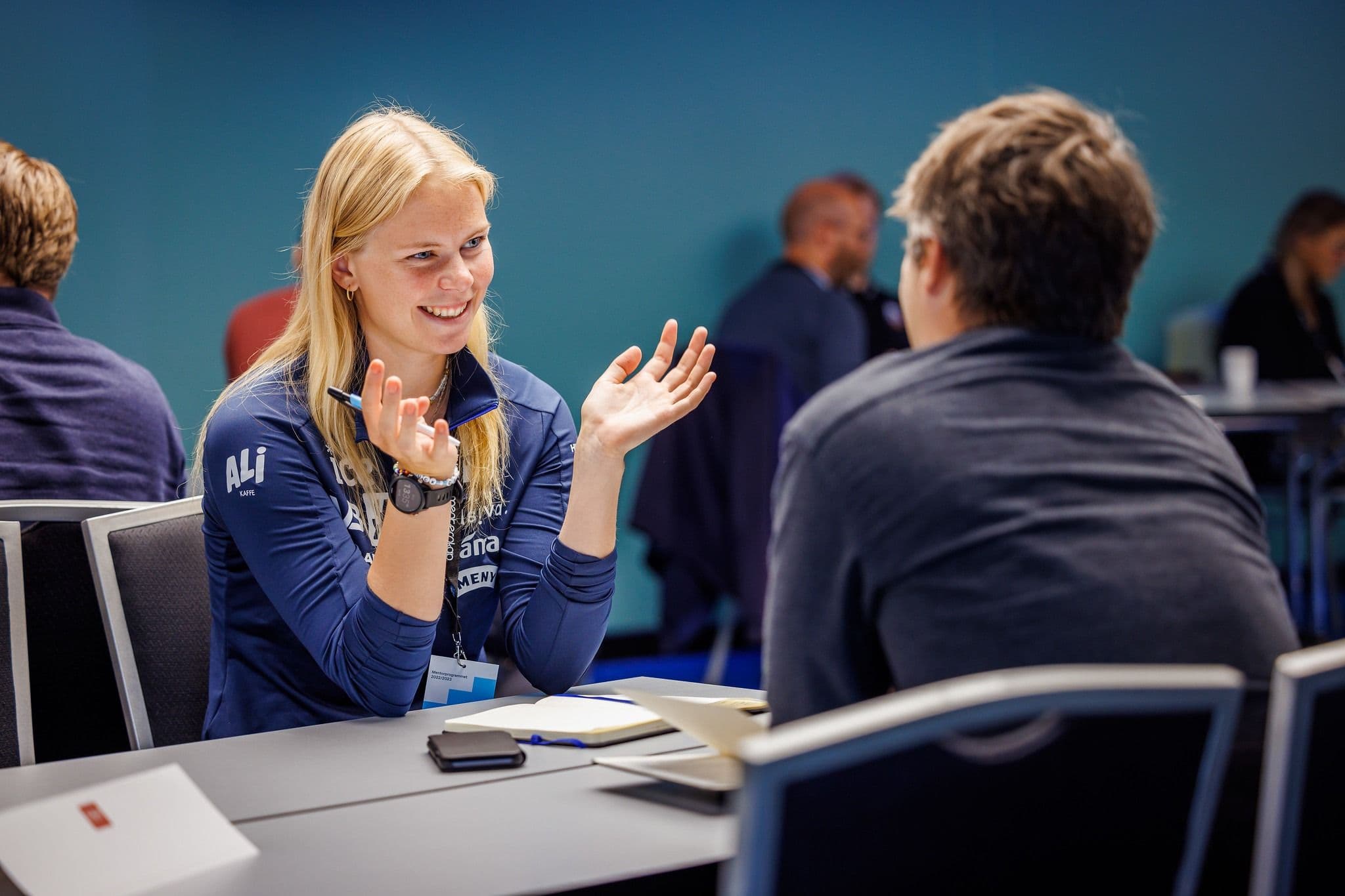 En kvinne gestikulerer engasjert i en samtale ved et bord på et seminar.