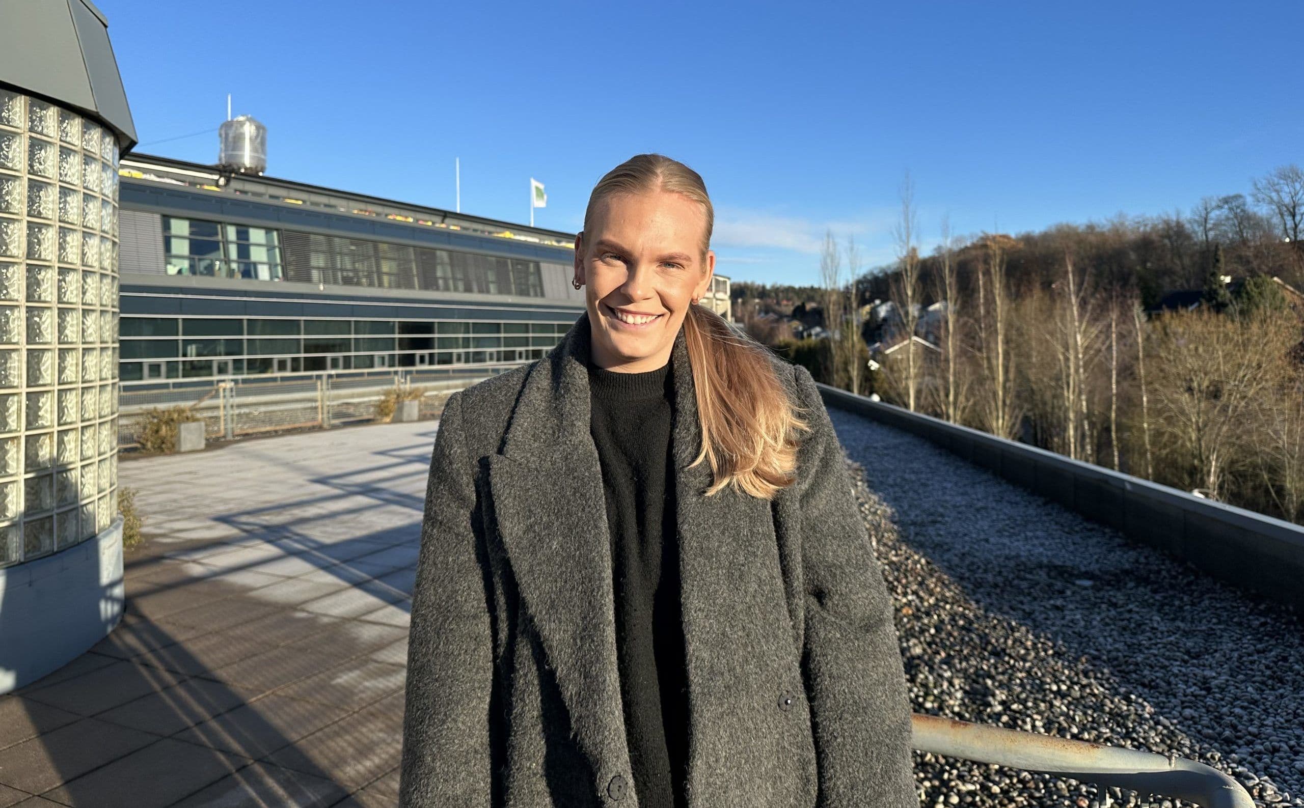 Portrett av en person i grå frakk på en takterrasse med en bygning i bakgrunnen.