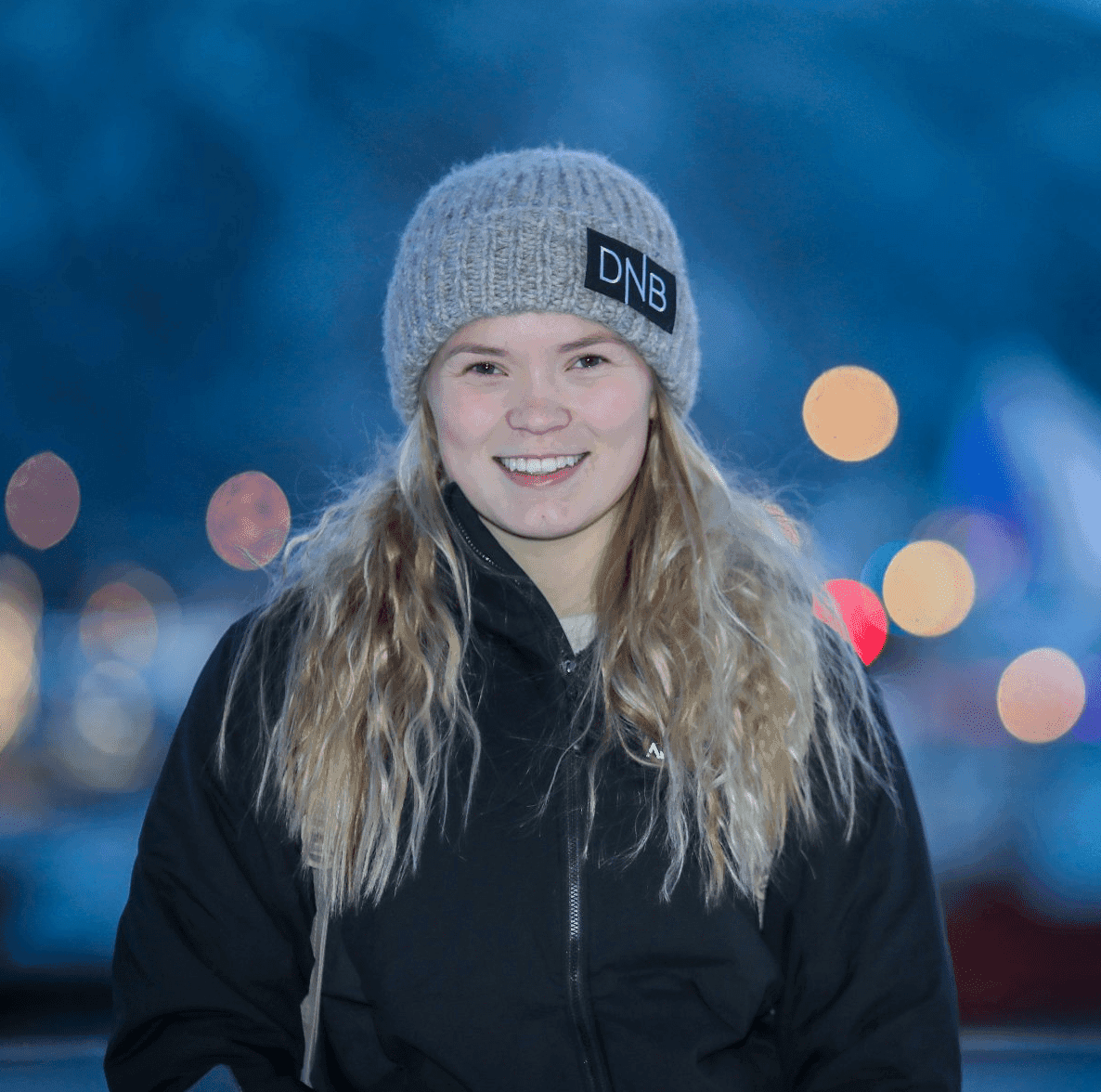 Portrett av en kvinne med grå lue og svart jakke med fargede lys i bakgrunnen.